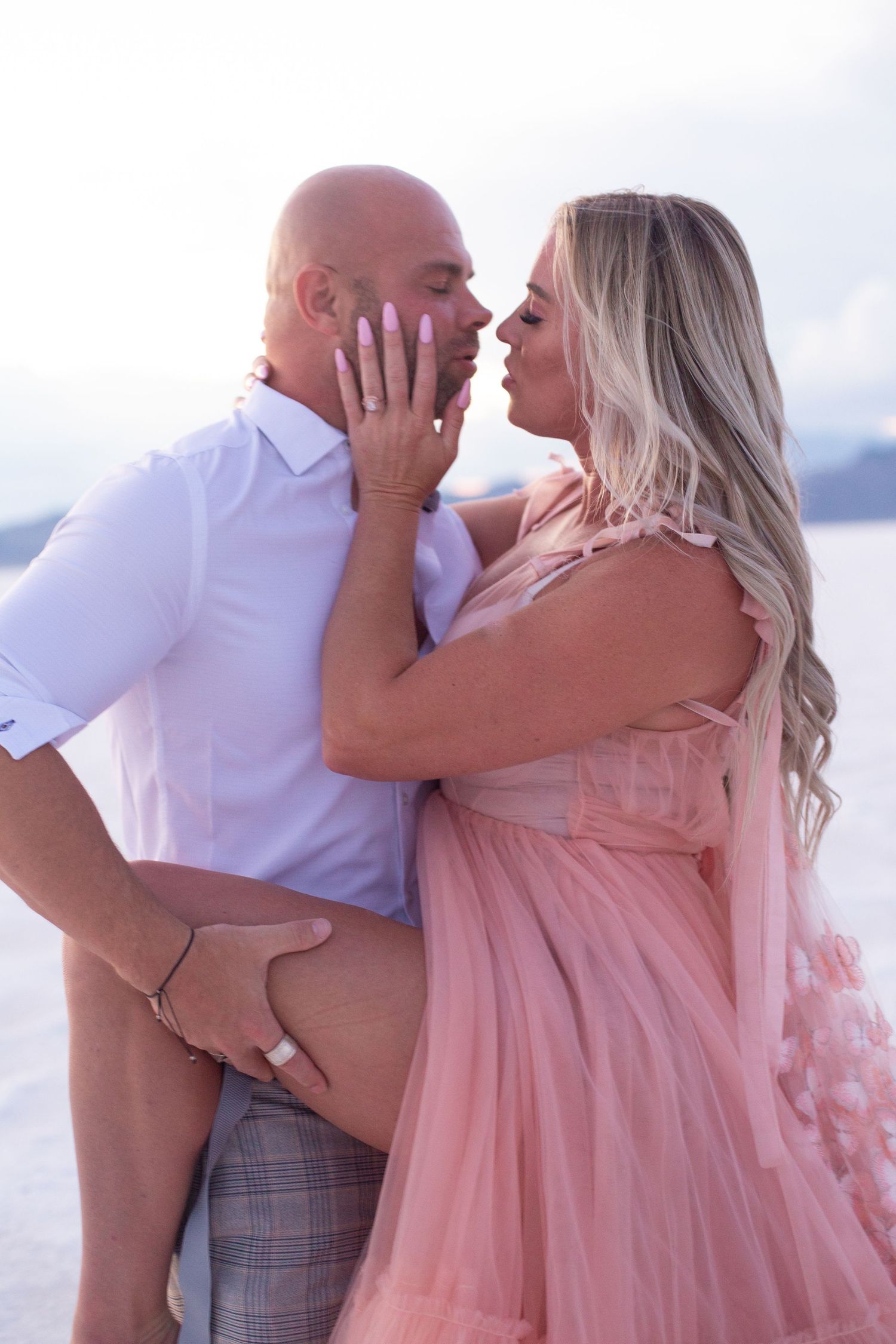 Romantic couple embraces on a beach at sunset with flowing pink dress creating ethereal atmosphere.