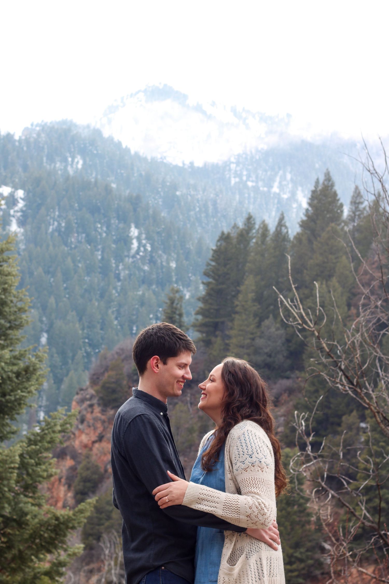 Couple sharing tender moment against backdrop of snow-covered mountain peaks and evergreen forest.
