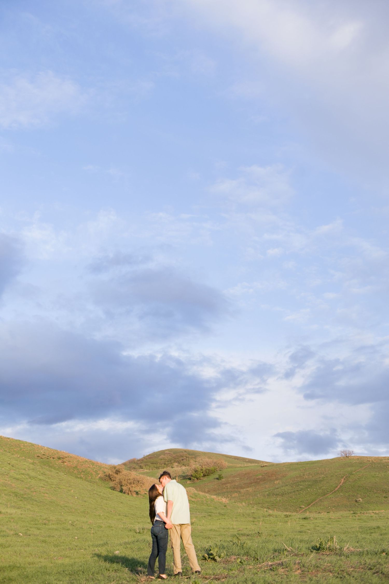 A couple embraces in a scenic field with rolling hills and blue sky in the background.