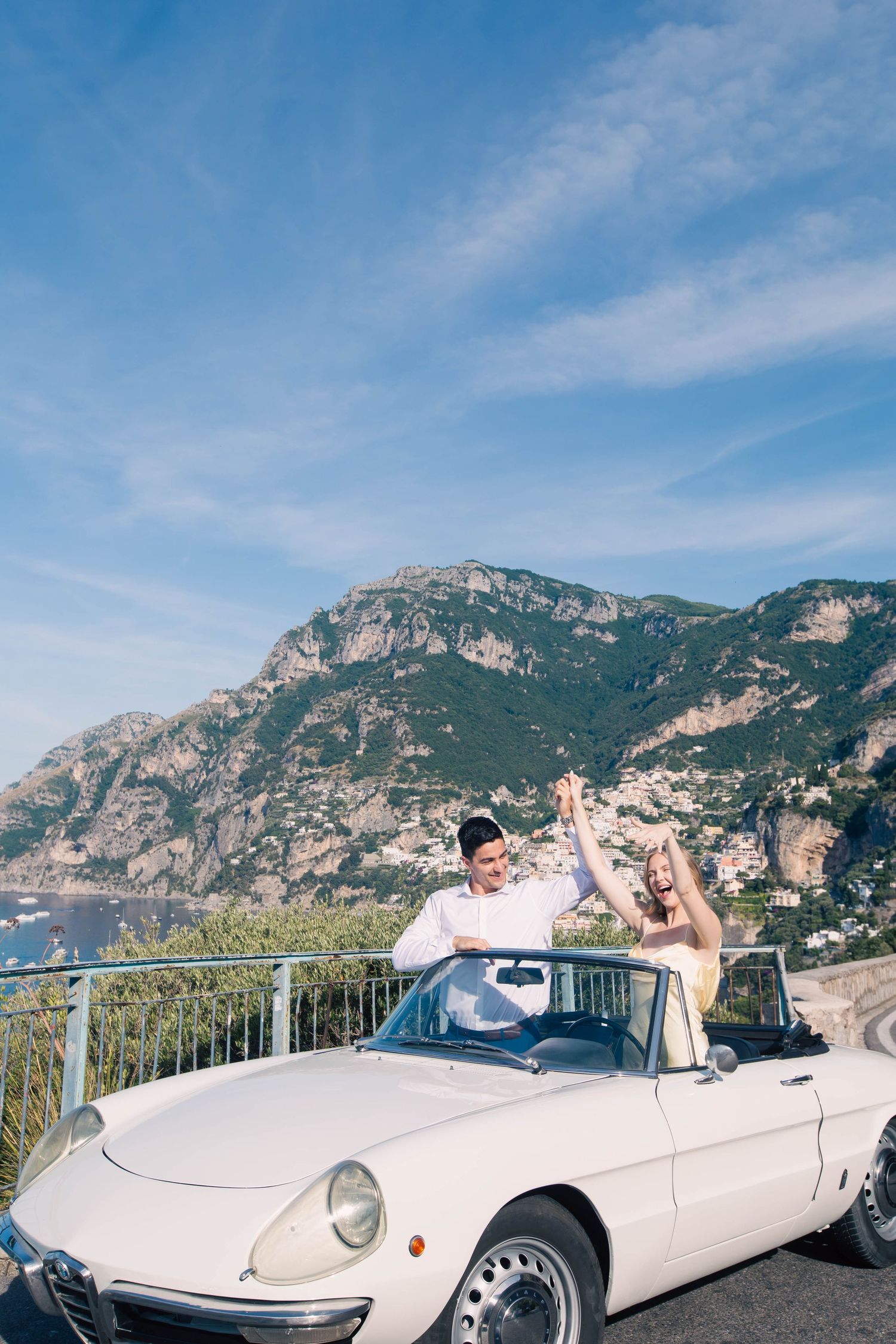 Couple celebrating in a white vintage convertible on the Amalfi Coast with scenic mountains and blue sky.