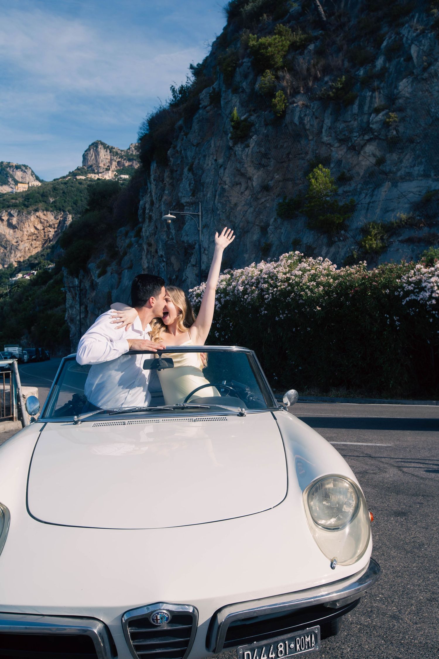 Joyful couple kissing in white vintage Alfa Romeo convertible with arm raised on Amalfi Coast.