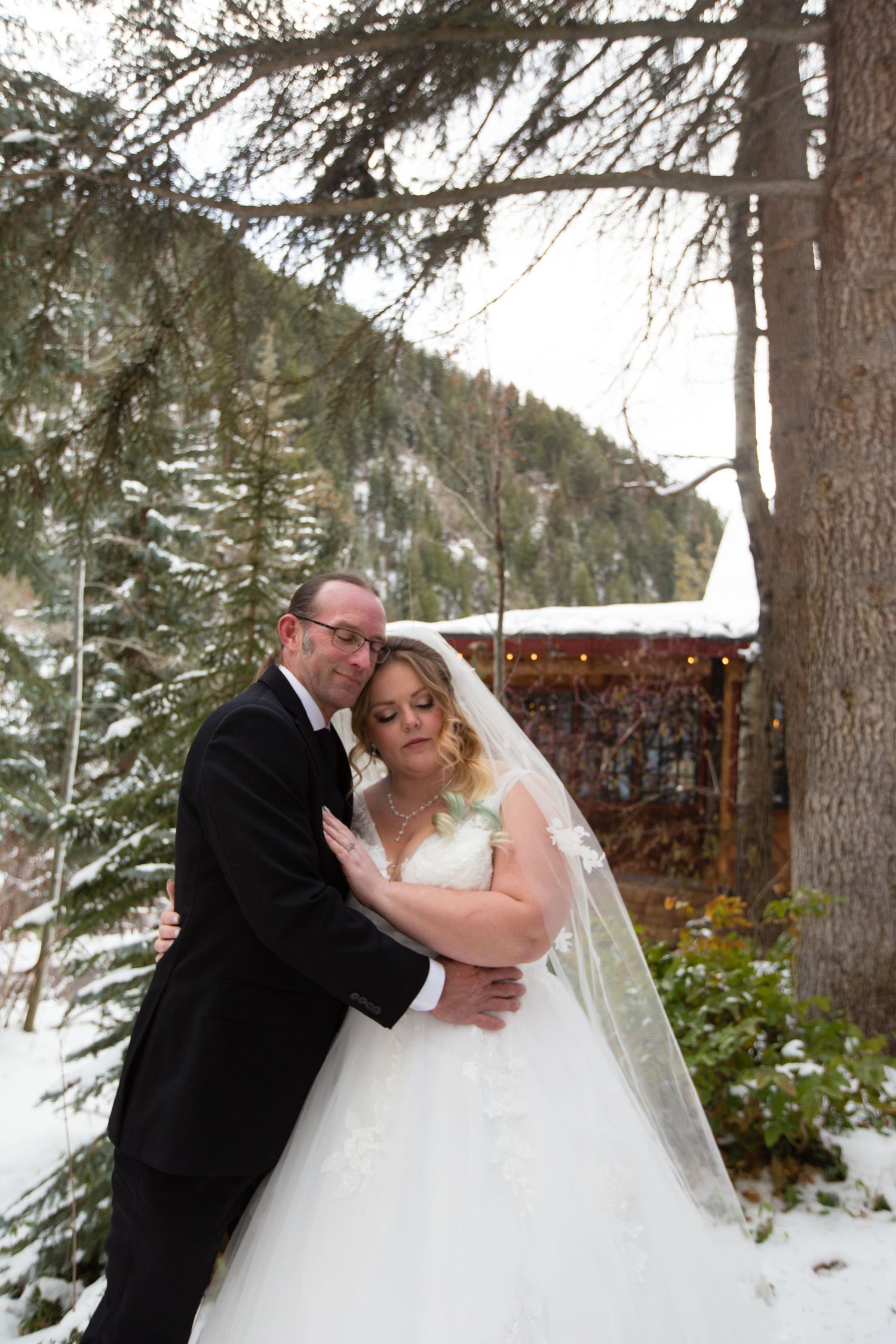 A romantic winter wedding photo captured in a snowy mountain setting with evergreen trees in the background.