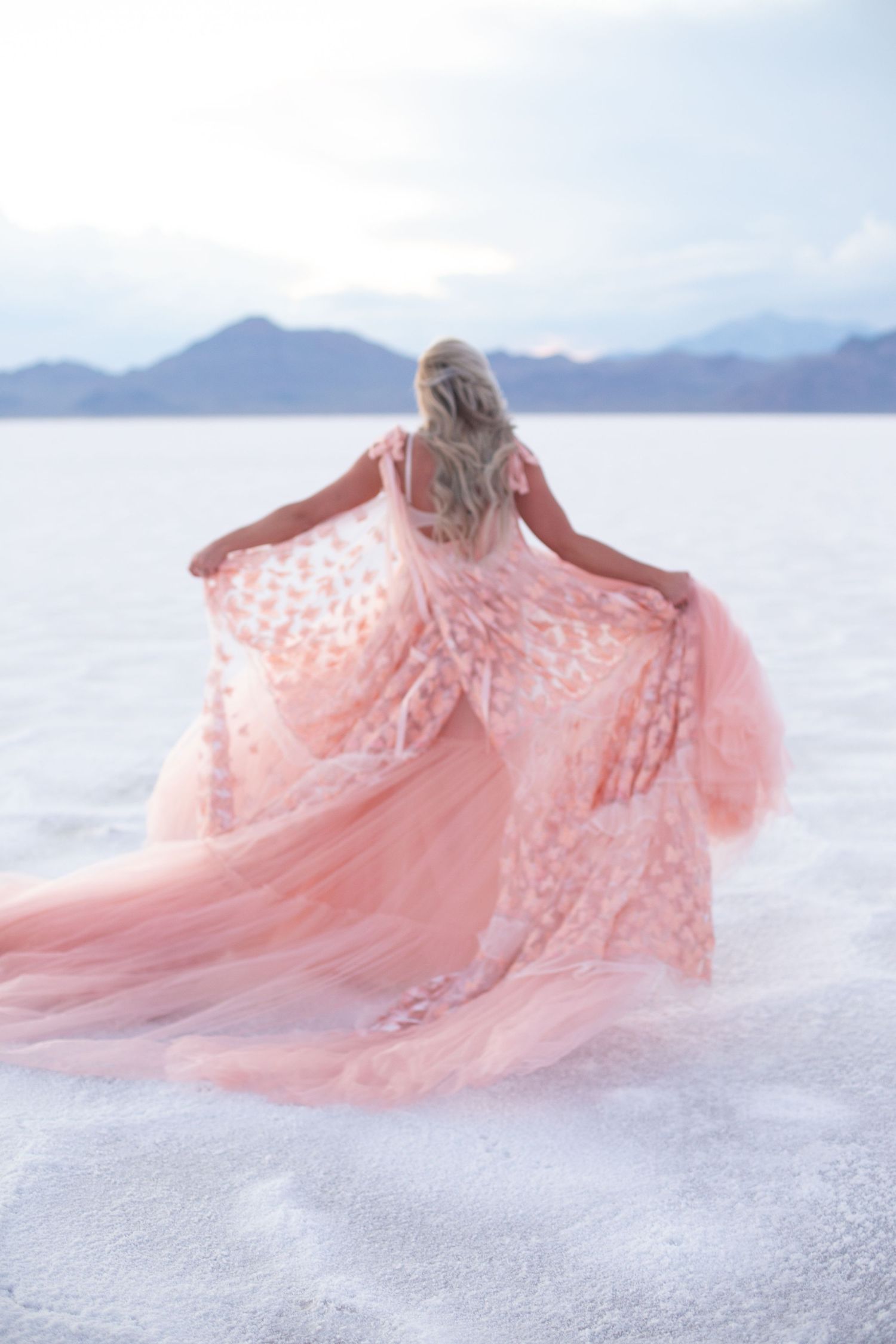 A pink lace dress spreads dramatically across white sand with mountains on the horizon.