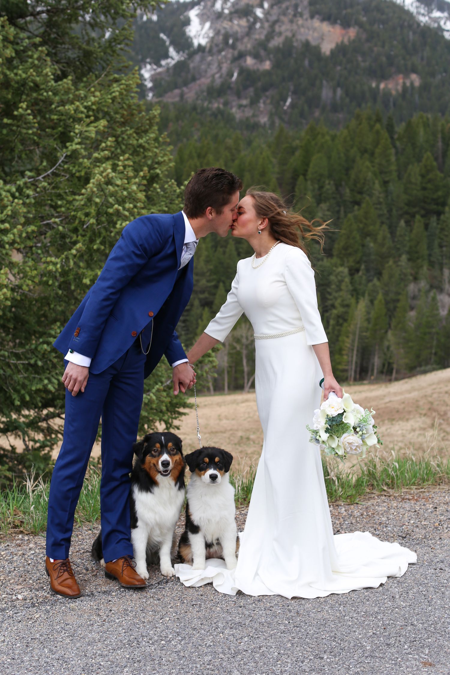 Couple with two dogs share romantic moment on mountain path during wedding photoshoot.