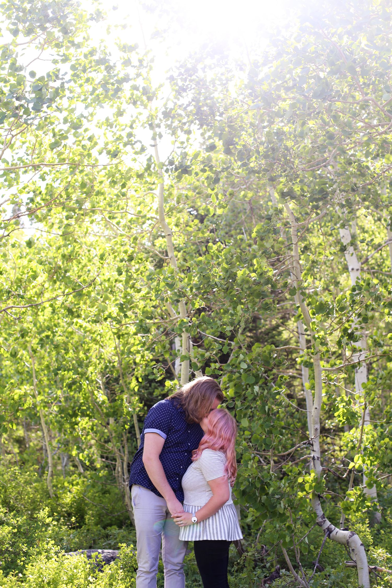 Couple kissing among sunlit green aspen trees in a forest setting, woman with pink hair wearing white top.