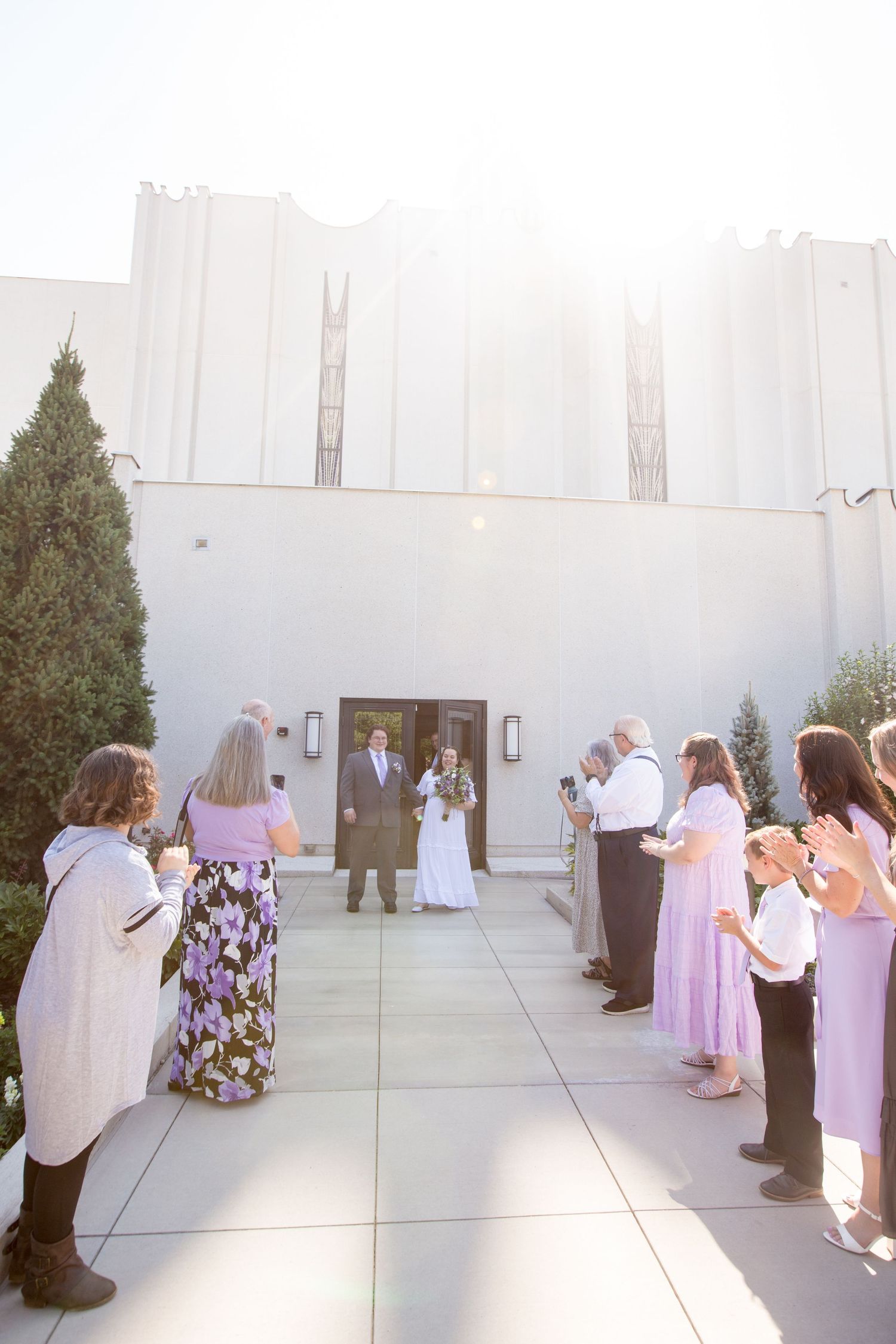 Wedding guests line a sunlit courtyard as a couple exits a white church building on their special day.