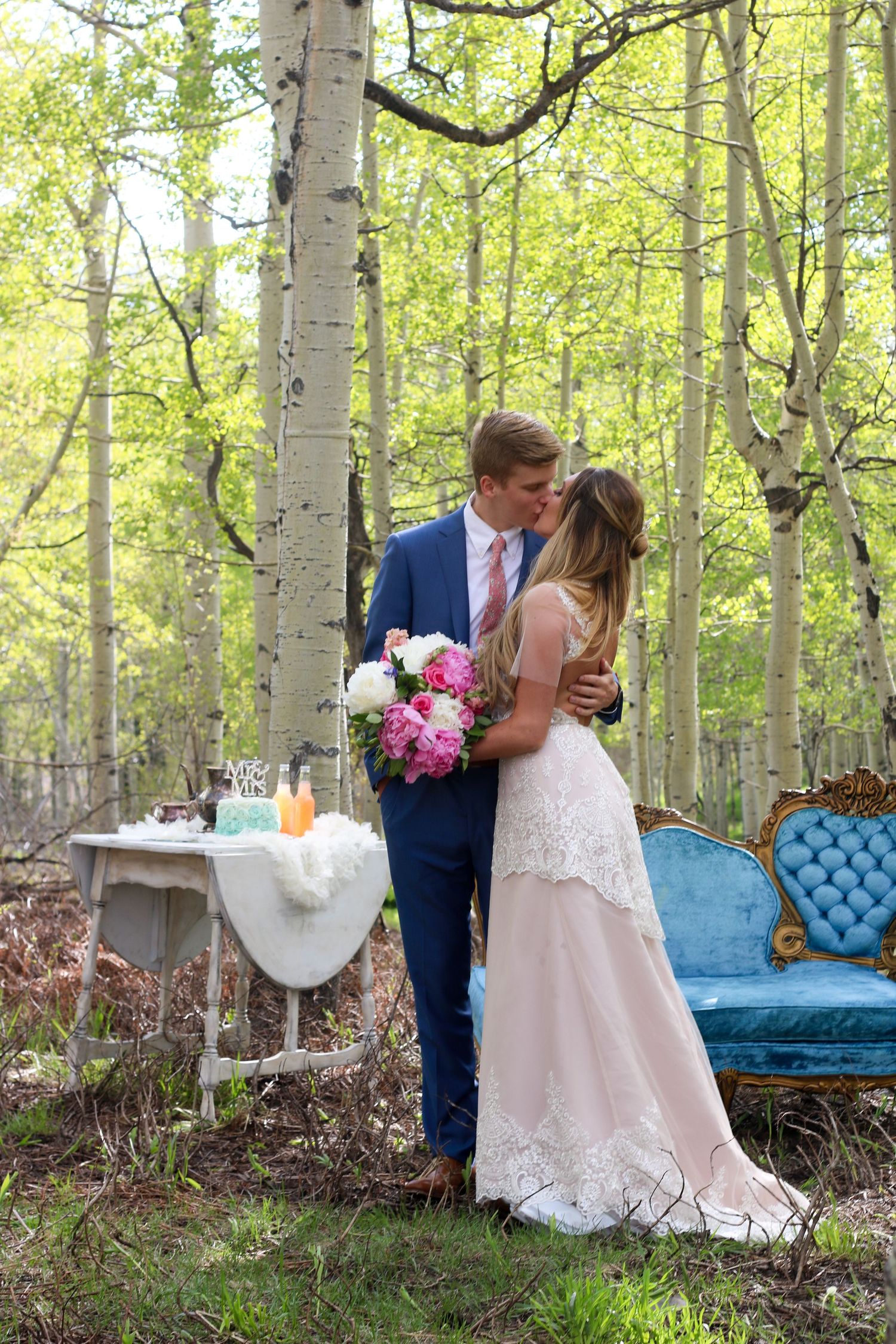 Bride and groom kissing in aspen forest, blue velvet sofa and floral bouquet, outdoor woodland wedding ceremony.