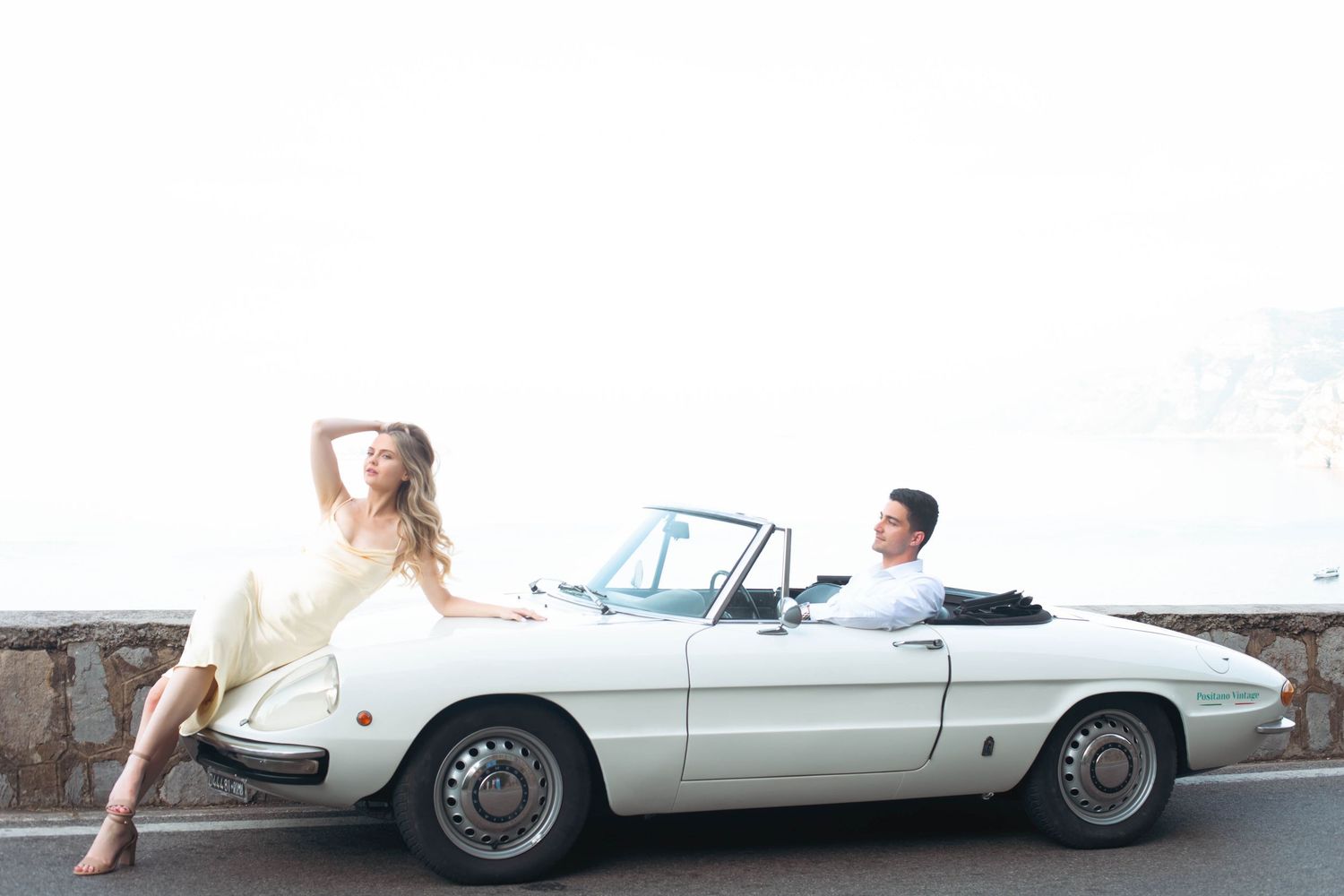 A woman in a yellow dress poses on a white vintage convertible sports car while a man sits in the driver seat.