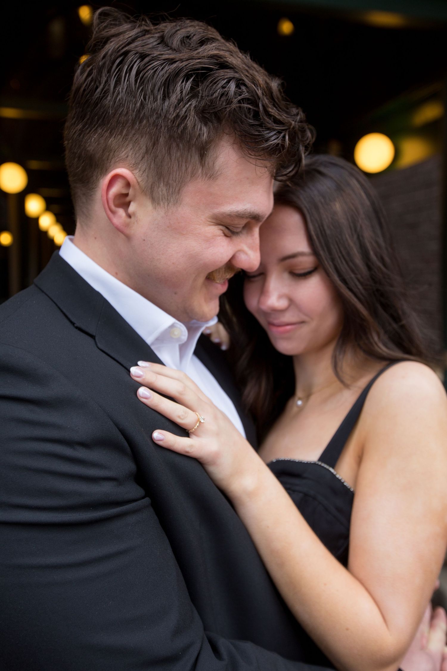 Romantic couple embracing closely, man in dark suit and woman in black dress, sharing an intimate moment with warm bokeh lights behind them.