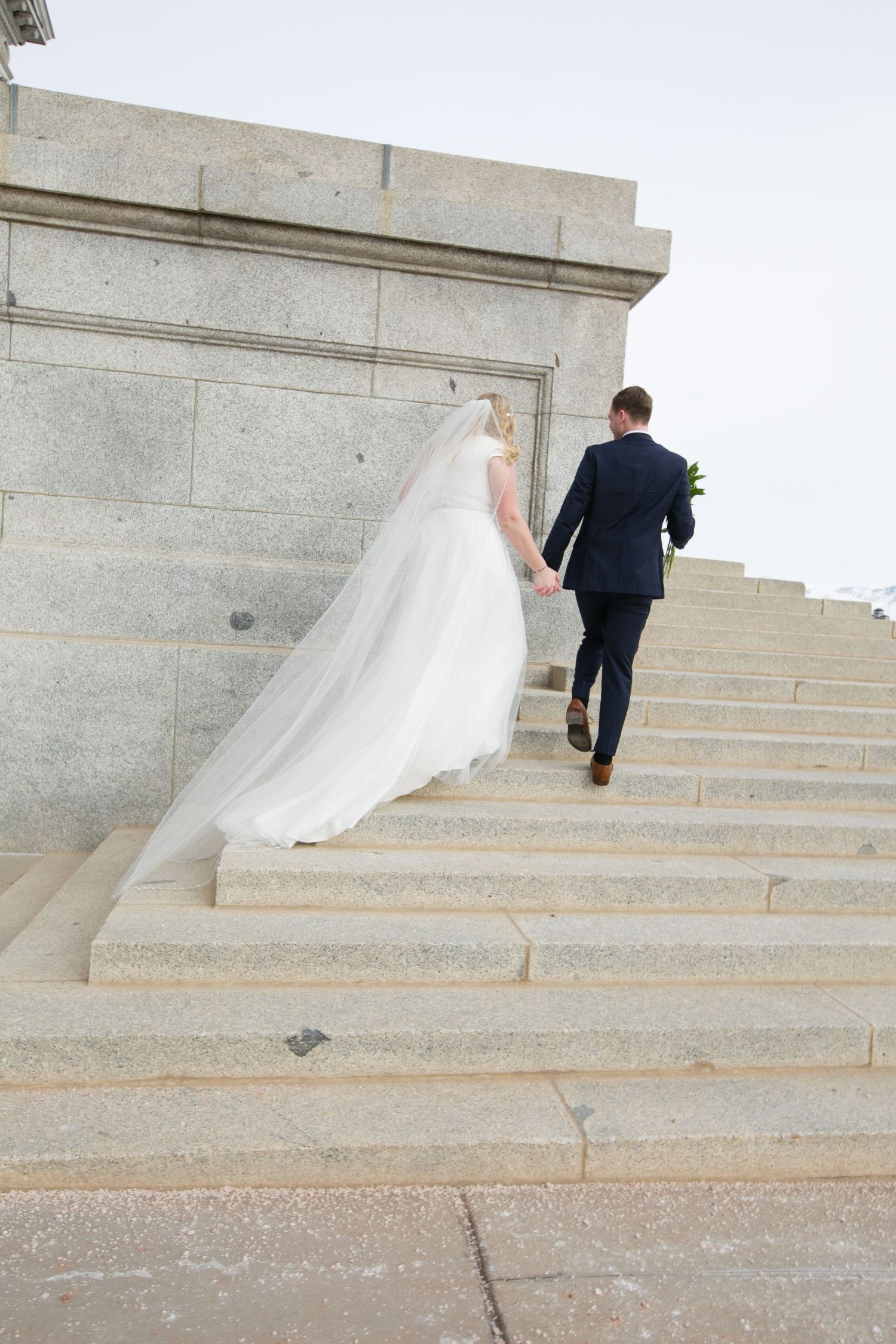 Bride and groom walk hand in hand up stone steps of a grand building, bride's veil trailing behind her.