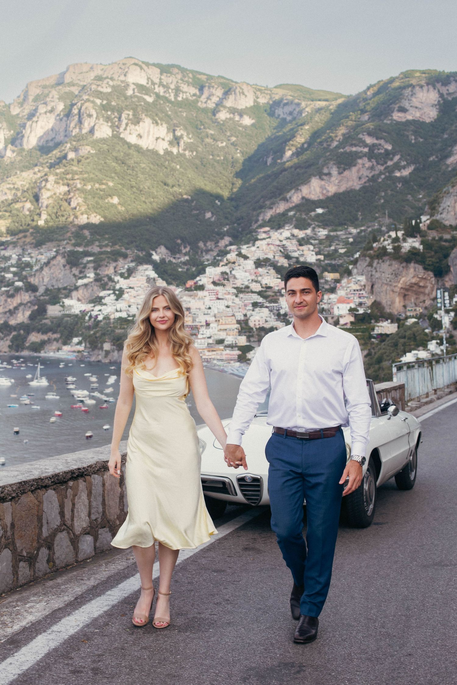 Elegant couple holding hands on Amalfi Coast road beside white vintage convertible with scenic backdrop.