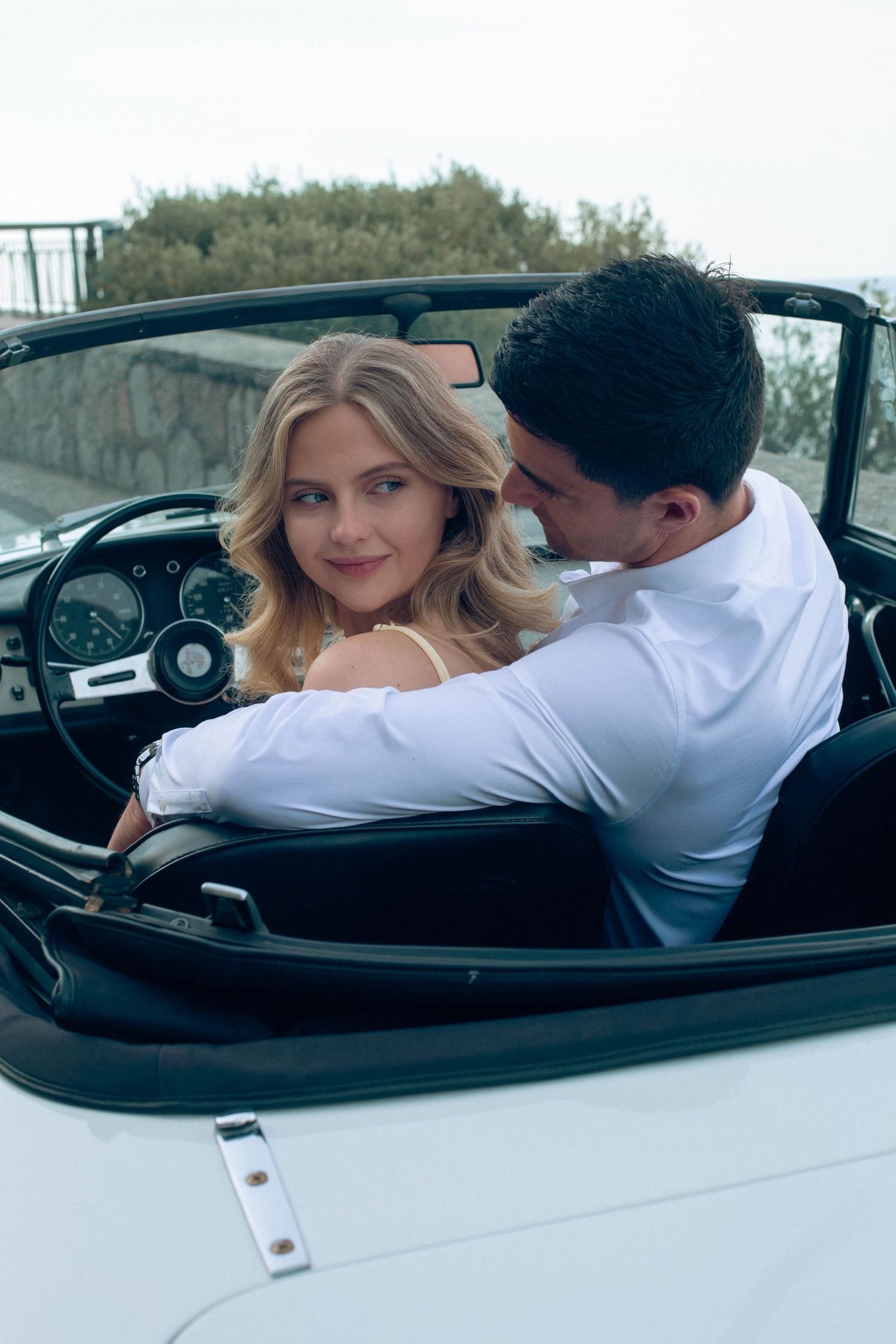 Blonde woman and man sharing romantic moment in classic white convertible car on Amalfi Coast.