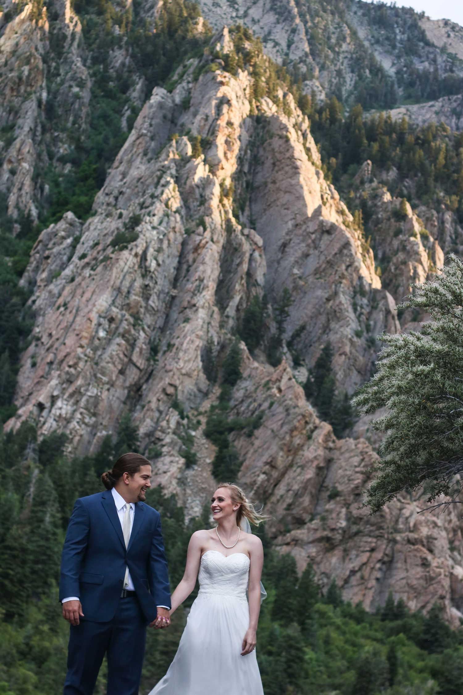 Couple in wedding attire posing against dramatic mountain cliffs during golden hour.