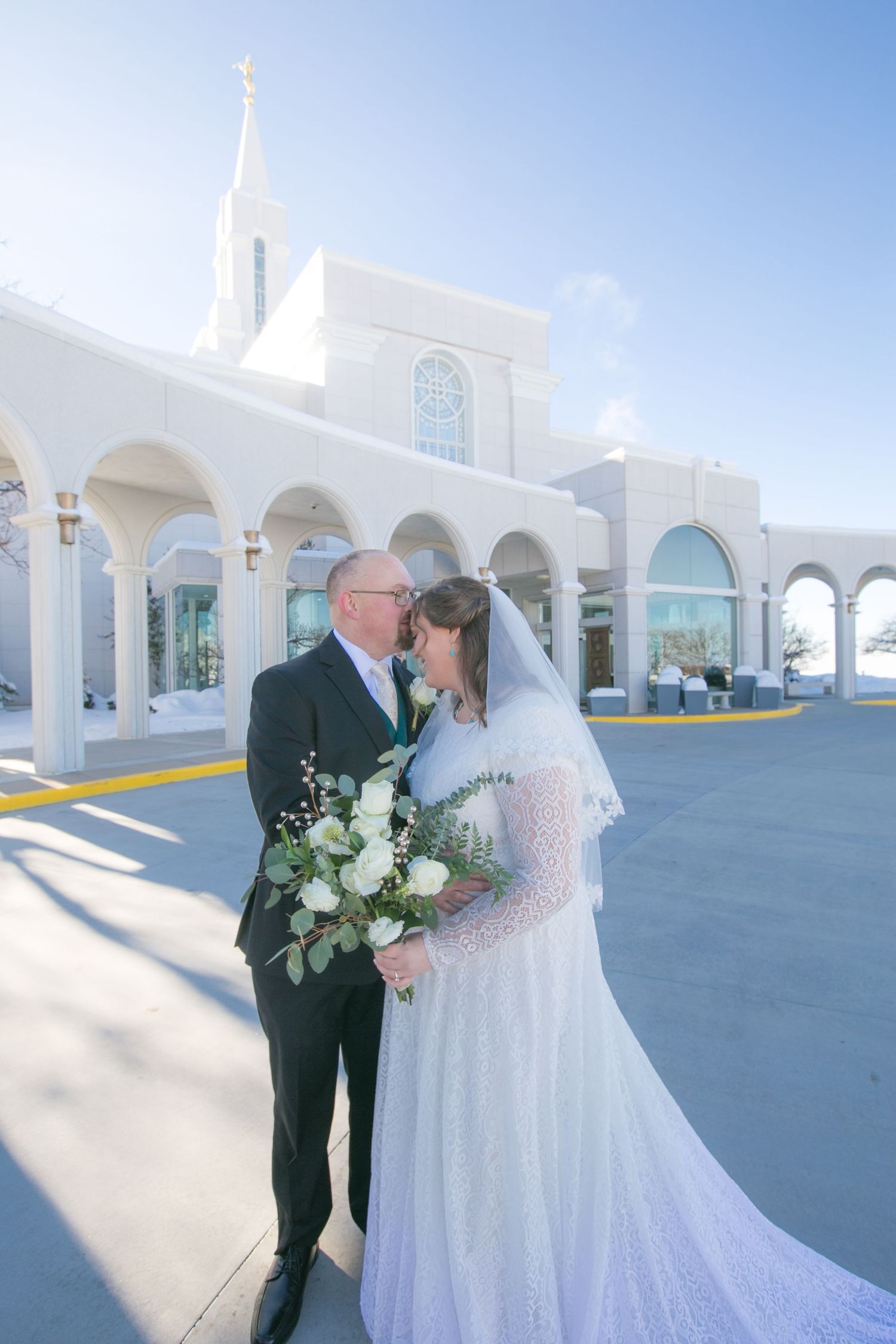Wedding couple poses in front of white church with elegant arches and steeple.