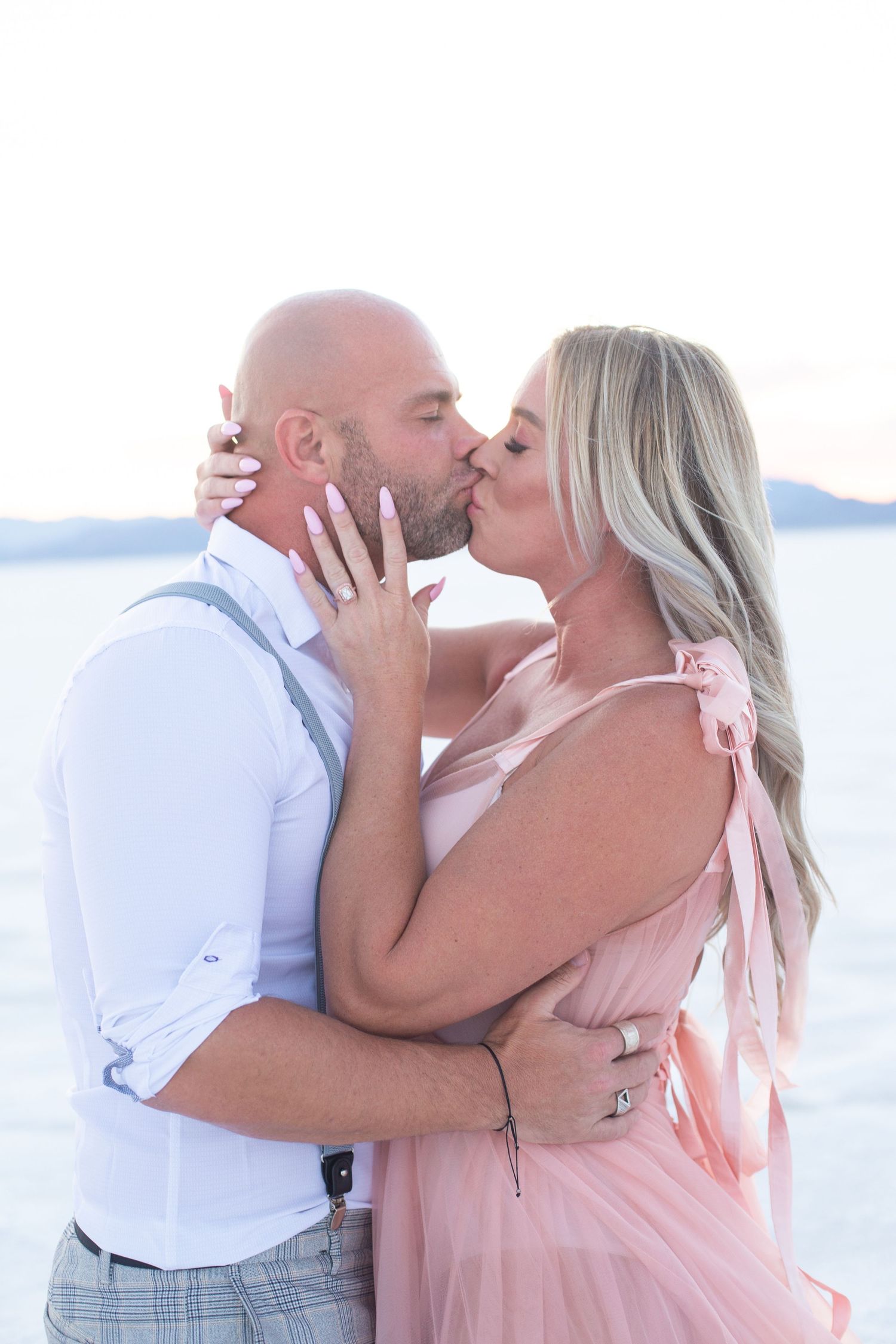 Tender moment captured between couple in pink dress and white shirt by ocean.