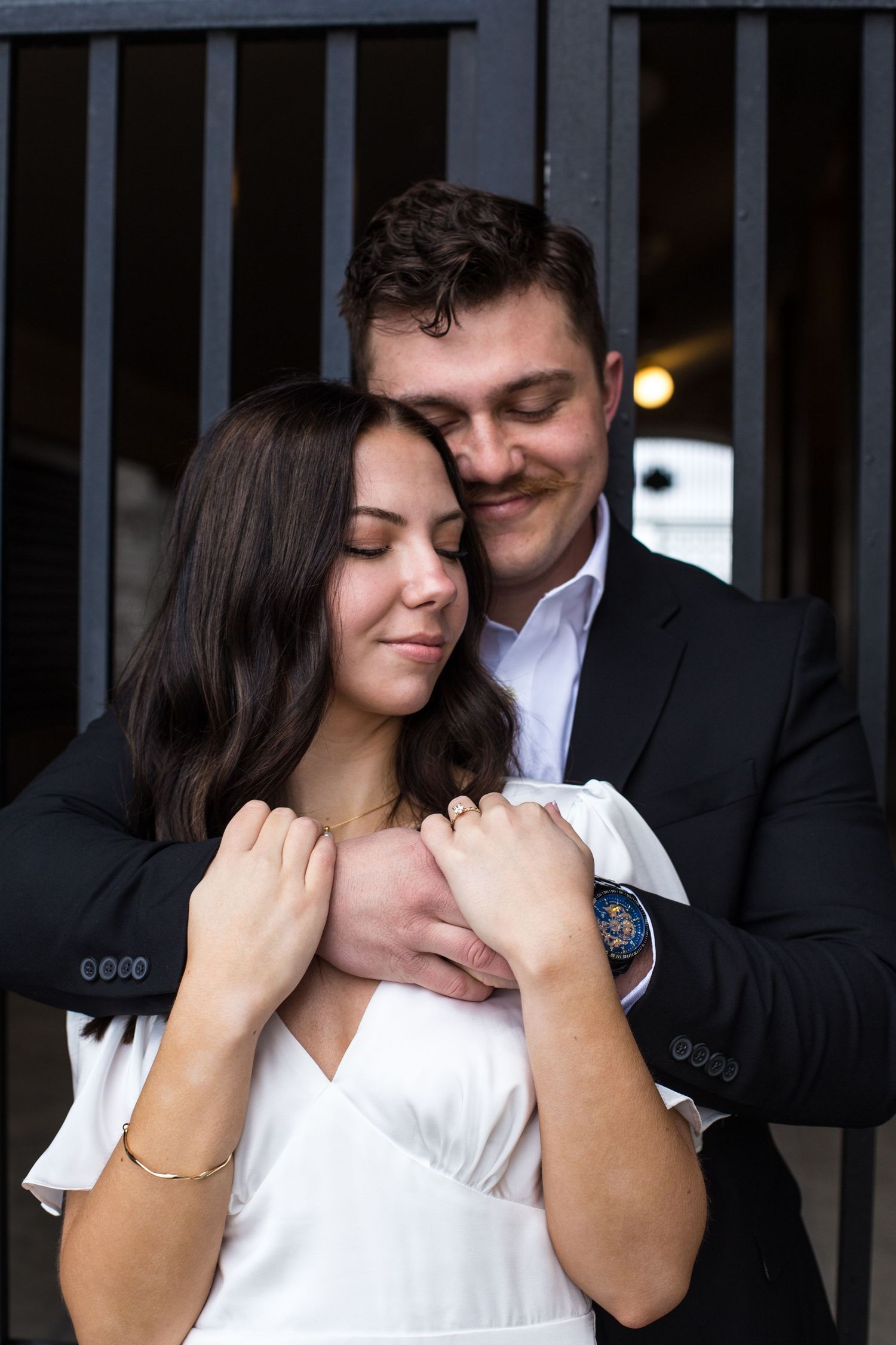 Romantic couple shares tender moments in formal attire against dark gates.