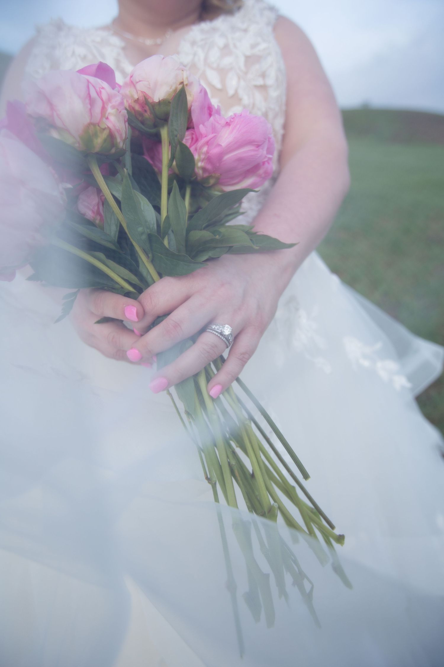 A delicate bouquet of pink roses held by hands wearing a white wedding dress and engagement ring.