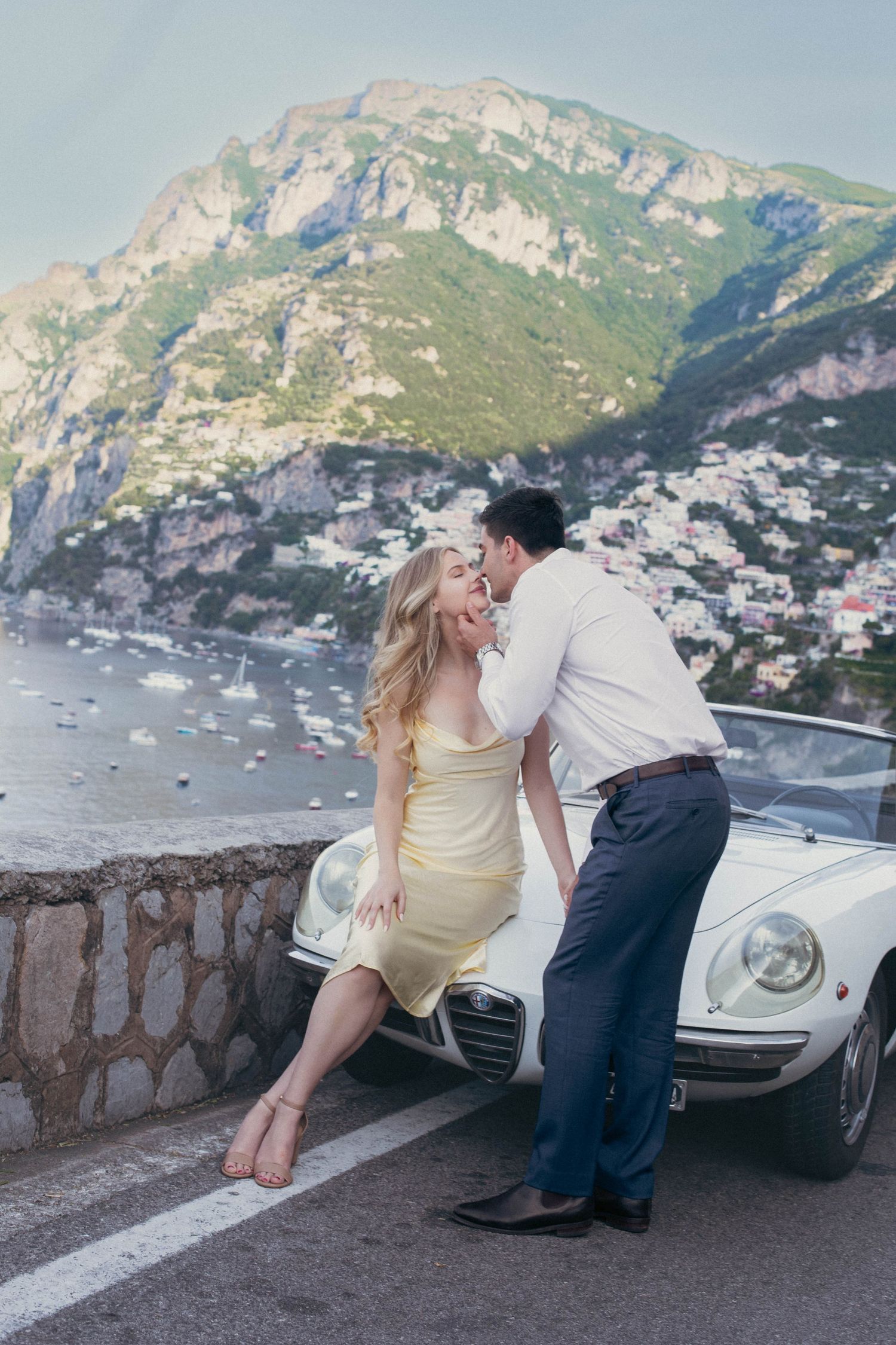 Laughing couple sharing a romantic moment leaning against a classic white Alfa Romeo Spider on the Amalfi Coast.