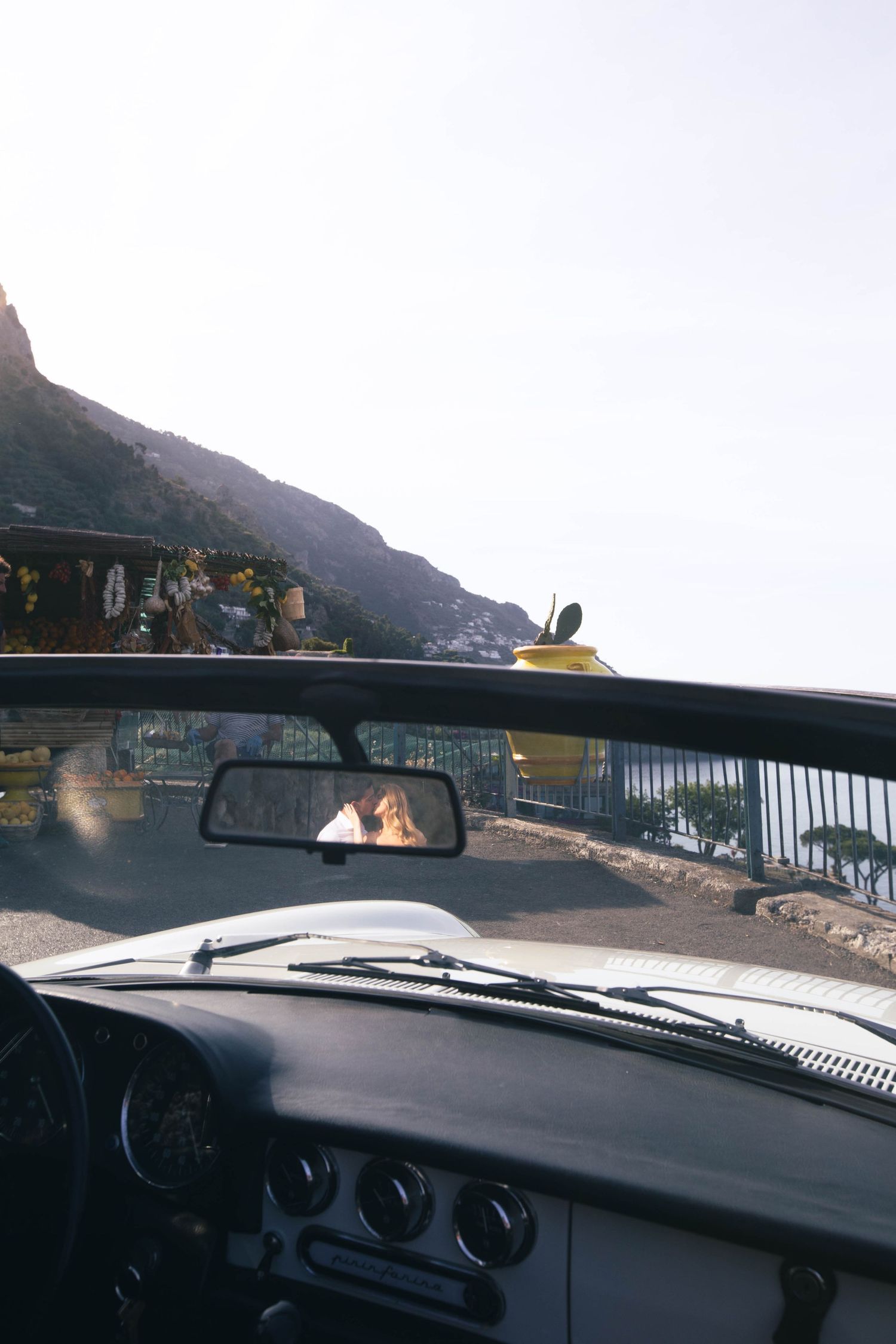 View from inside a vintage convertible car showing couple kissing in rearview mirror on scenic Amalfi Coast road.