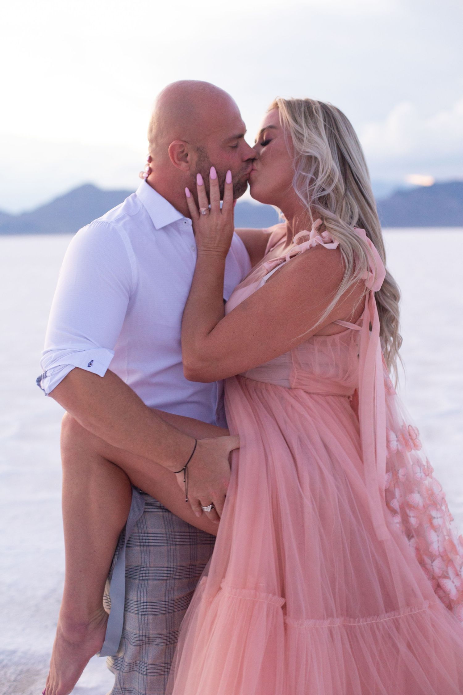Intimate couple's portrait with flowing pink dress against mountainous landscape.