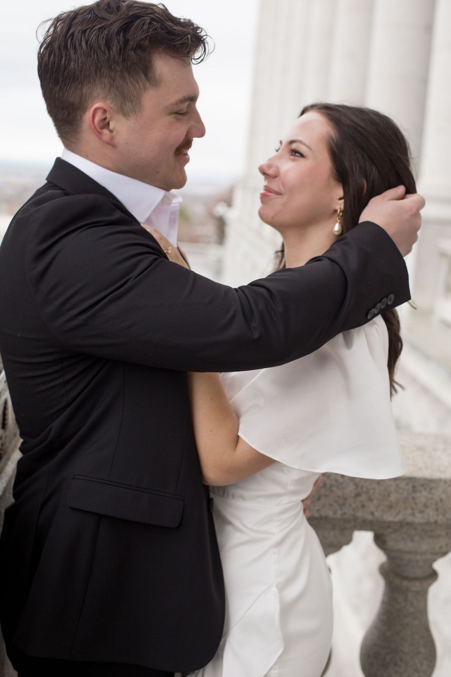A joyful embrace between a couple in wedding attire on a classical stone balcony.