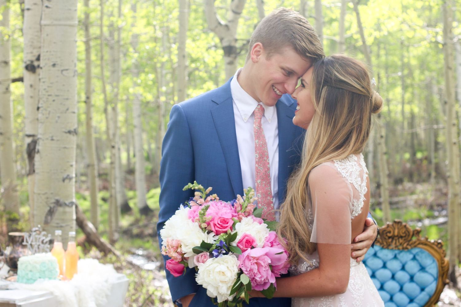 Wedding couple sharing tender moments in sunlit aspen grove with decorative blue seating and floral accents.