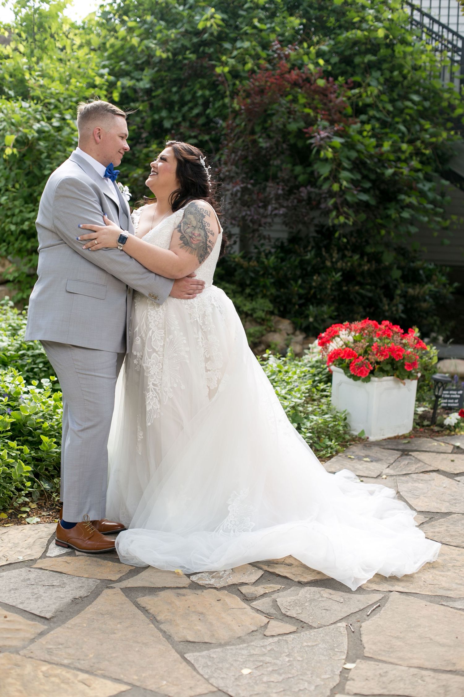 A bride and groom embrace on a stone patio surrounded by lush greenery and red flowers on their wedding day.