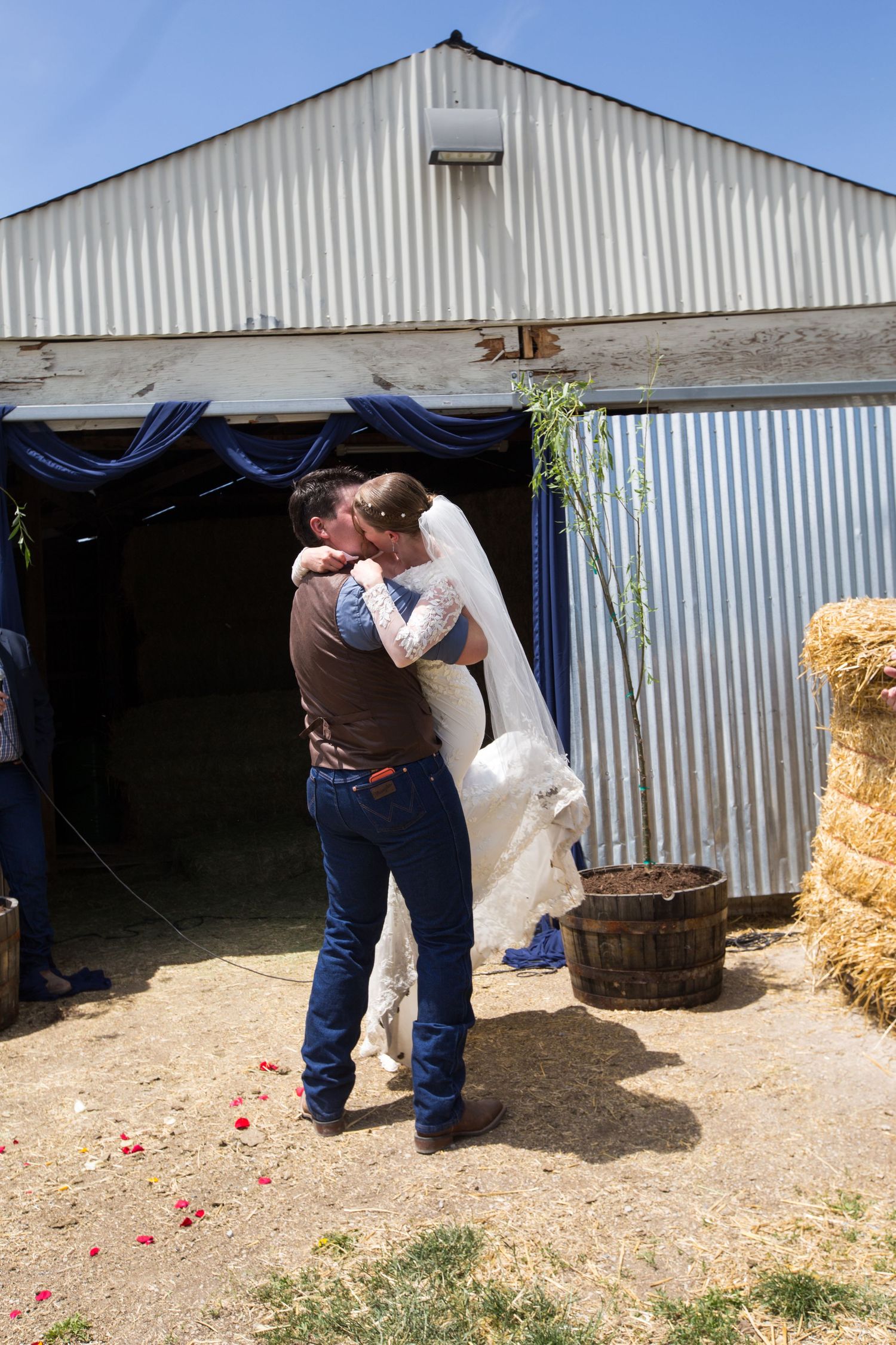 Bride and groom share first kiss outside rustic barn wedding venue with hay bales and blue draping decorations.