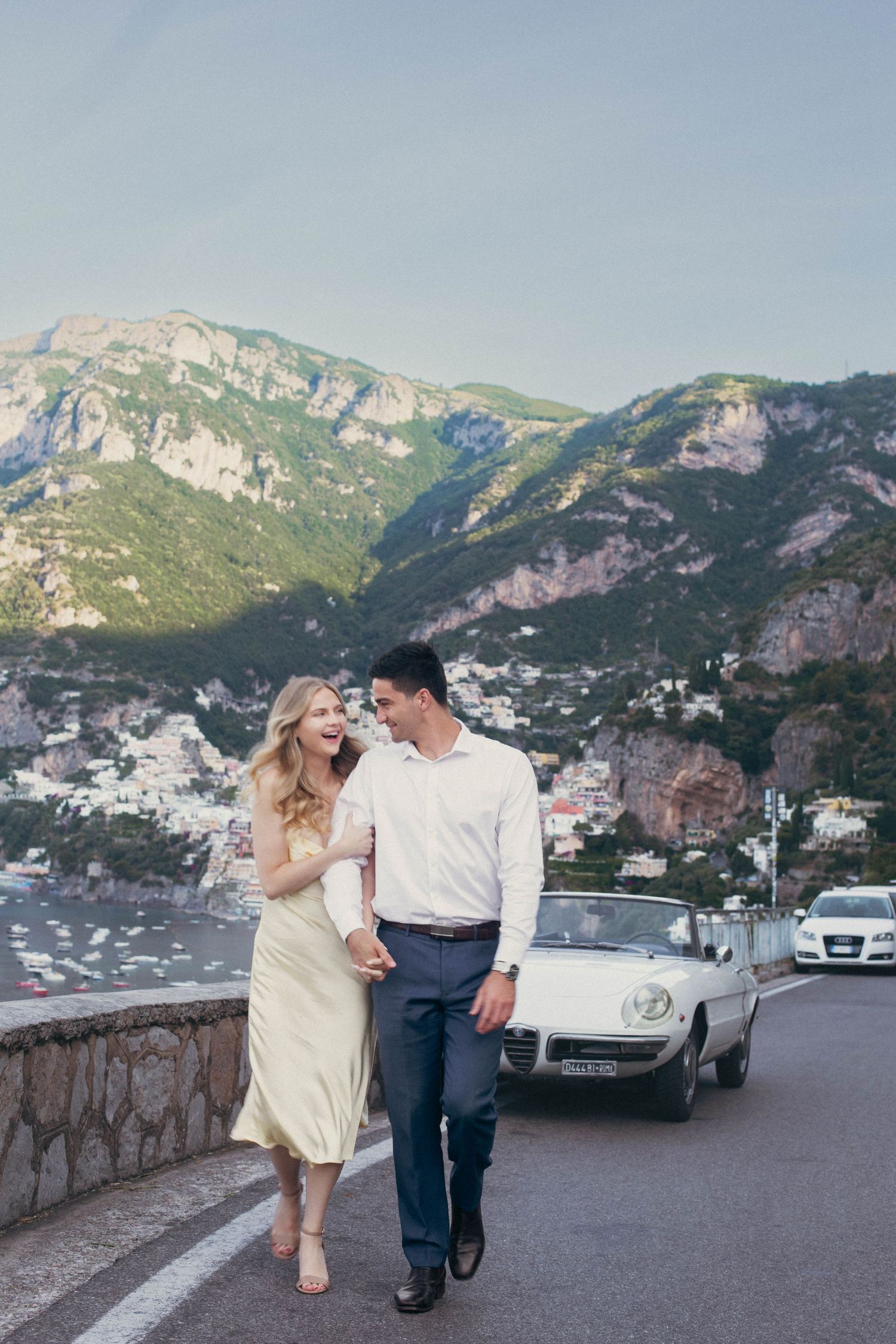 Couple walking hand in hand on Amalfi Coast road with vintage white convertible and Positano cliffside village backdrop.