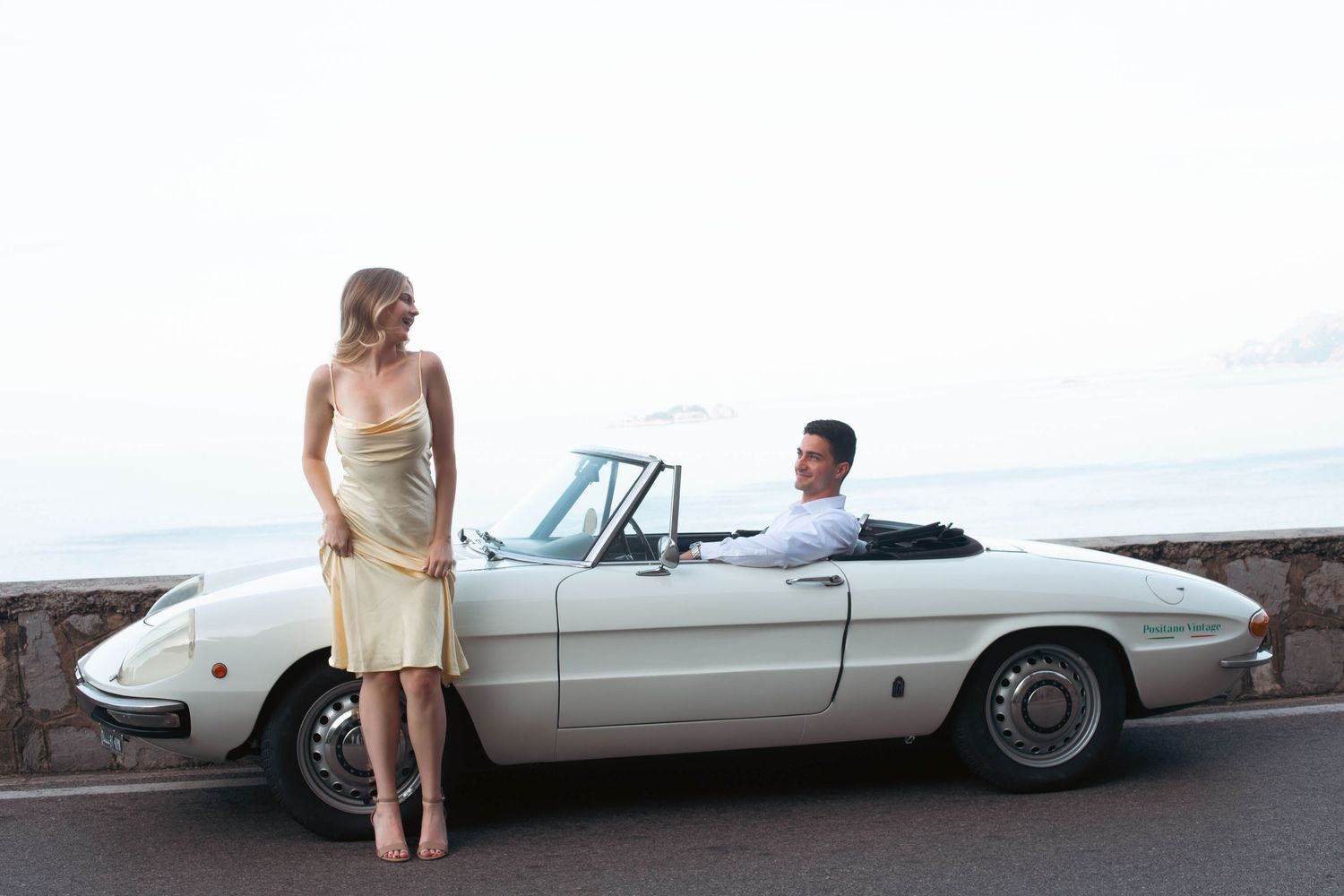 Woman in gold dress standing beside vintage white Alfa Romeo Spider convertible on Amalfi Coast overlook.