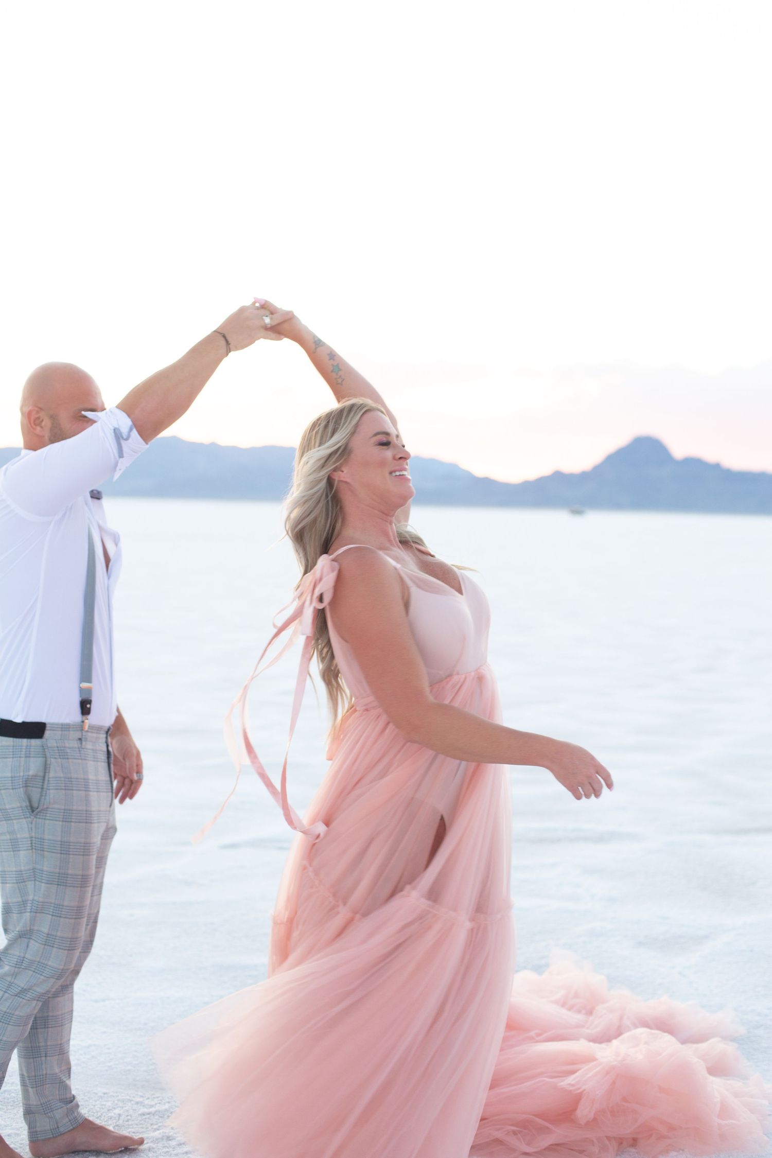 Dancing couple in formal attire twirl on salt flats with mountains in the distance.