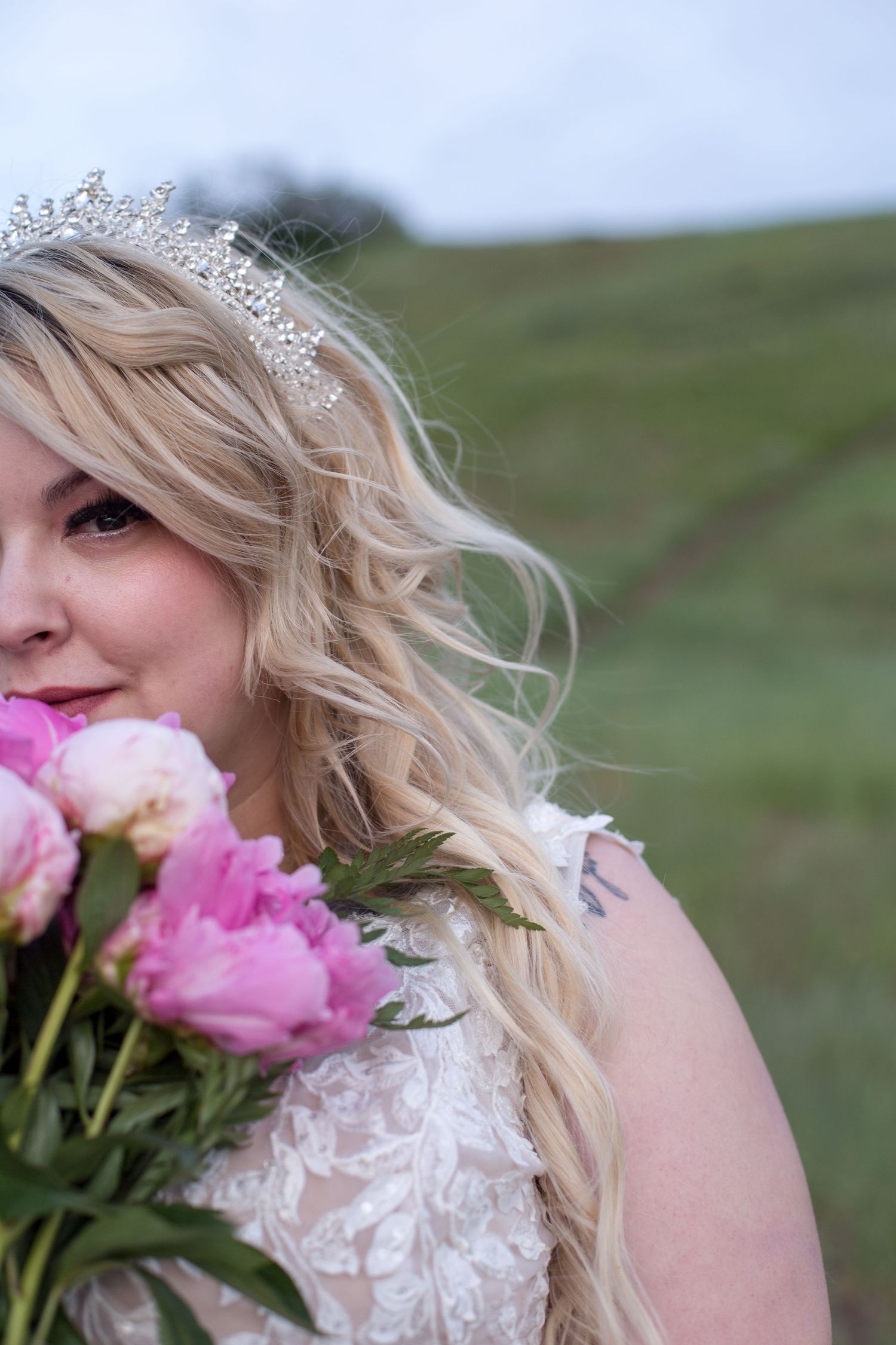 A bride wearing a crystal tiara holds pink peonies against a pastoral hillside background.