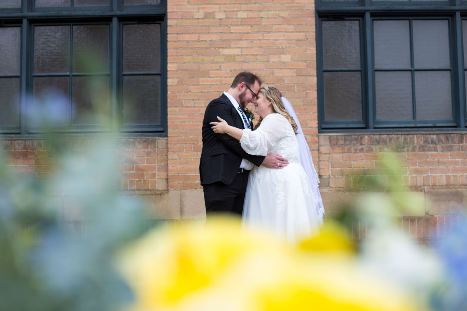 Romantic moment captured through flowers with brick building in background.