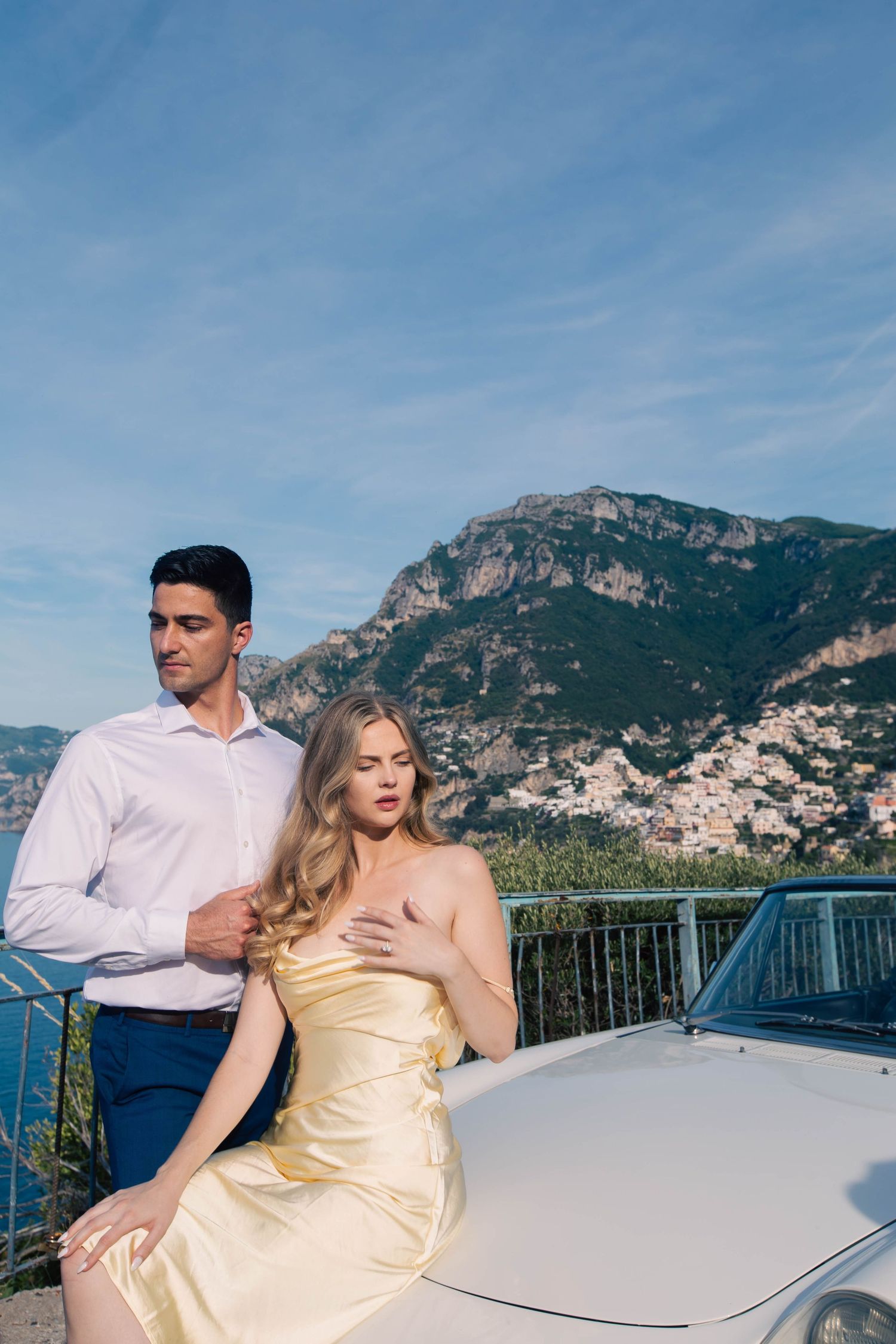 Elegant couple leaning on a vintage white car with stunning Positano cliffside village in the background.