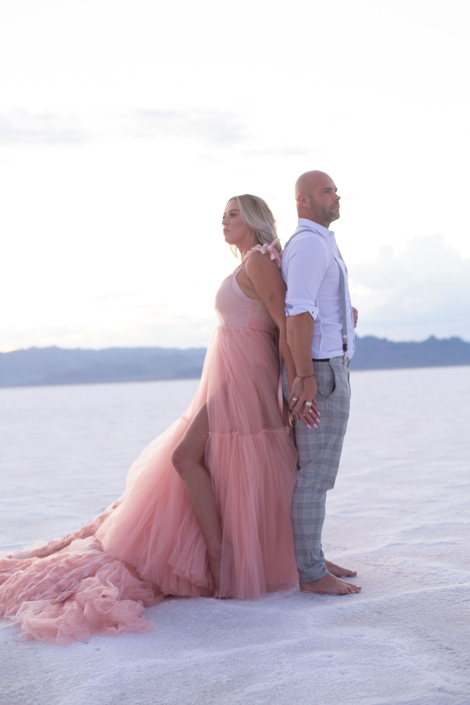 Romantic couple sits together on white sand wearing pink gown and formal attire.