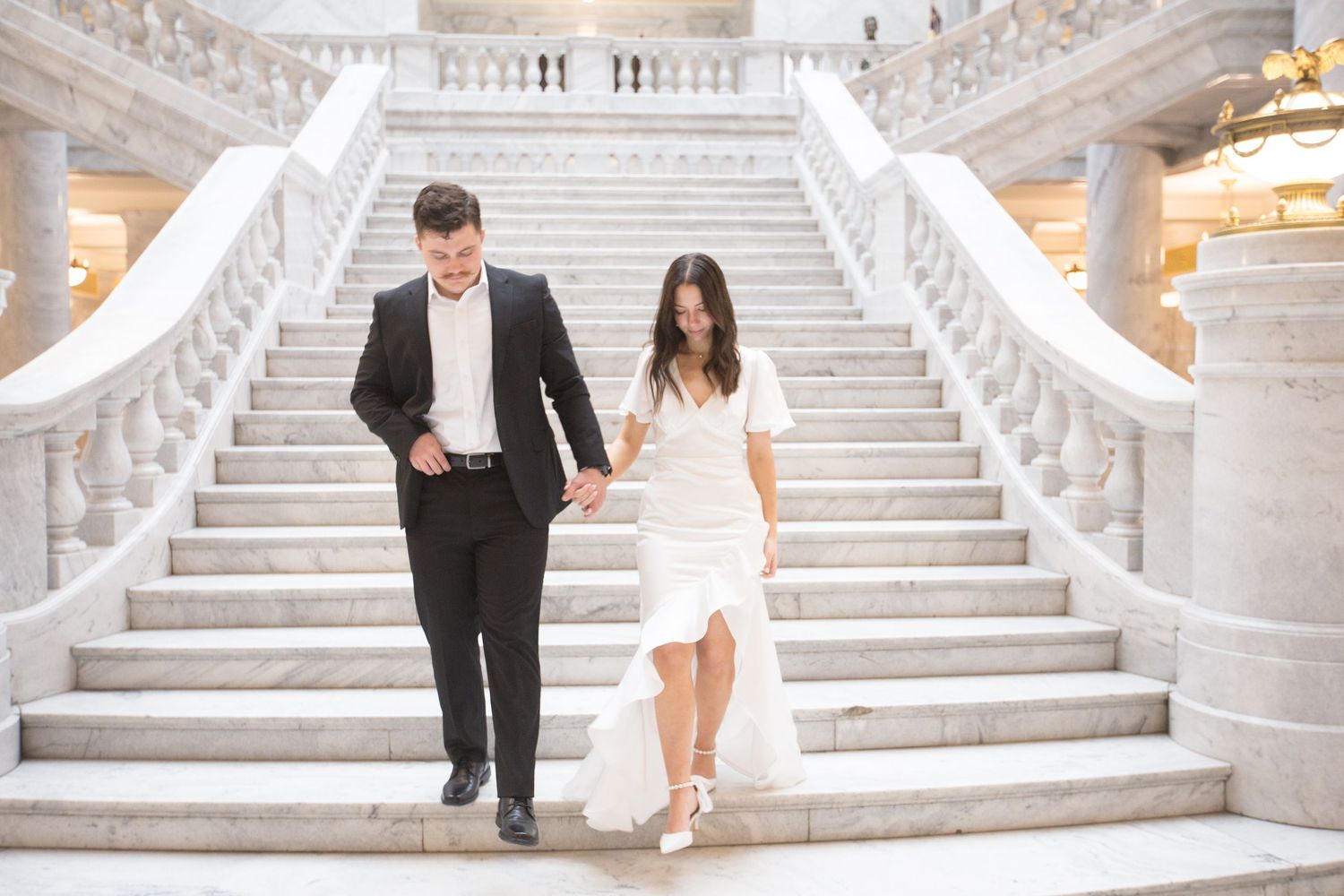 A couple descends grand marble stairs beneath classical architecture and painted murals.