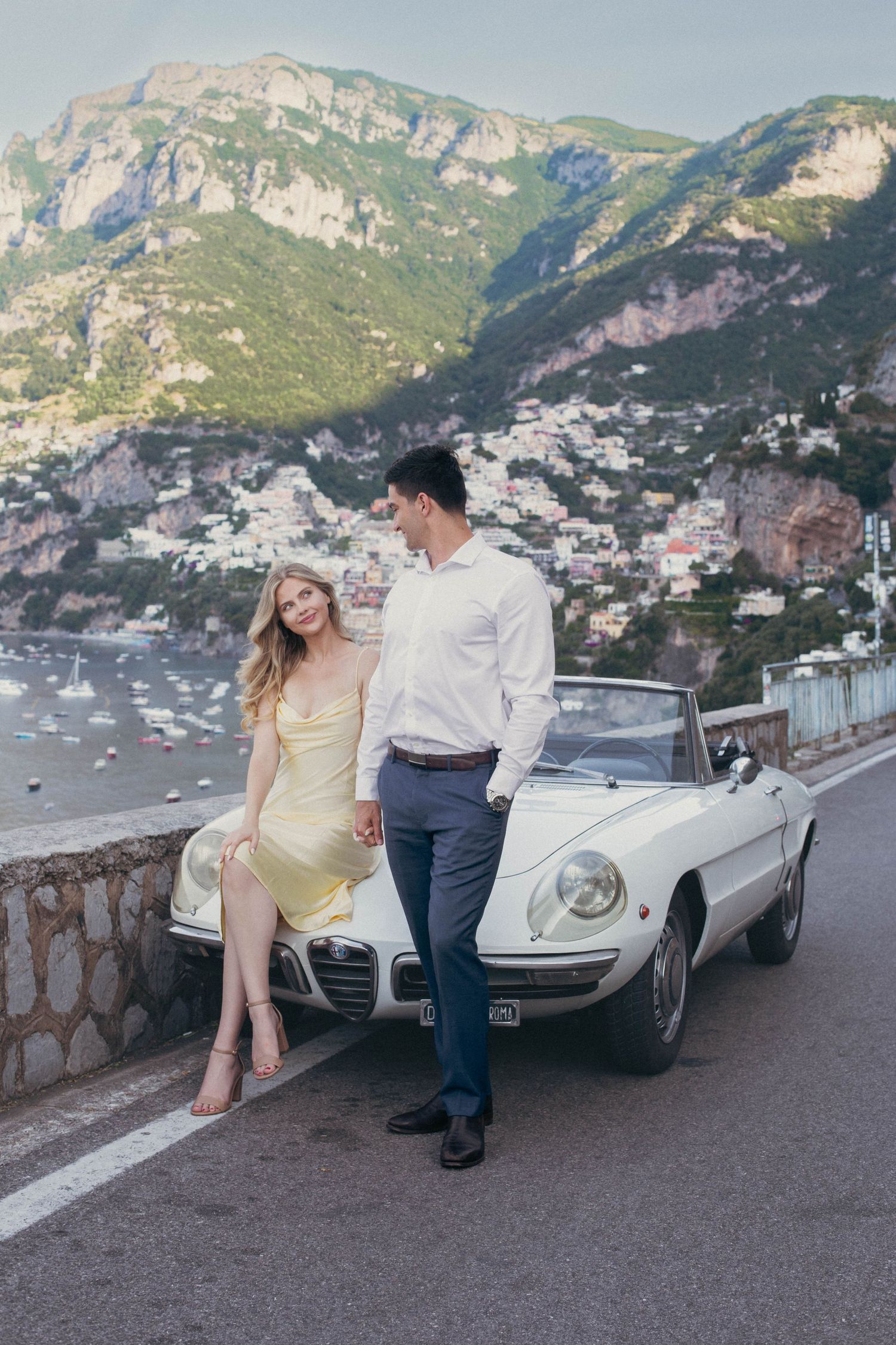 Stylish couple posing beside white Alfa Romeo Spider convertible with stunning Positano cliffside backdrop.