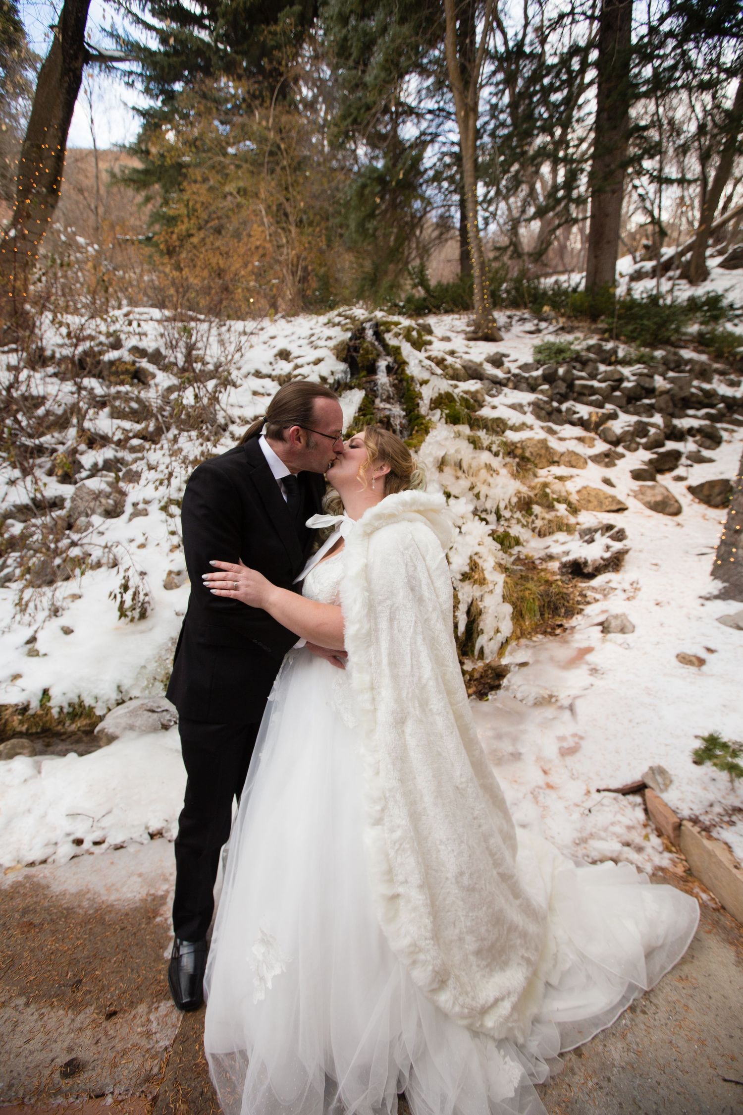 Romantic winter wedding portraits with couple embracing on snowy path surrounded by trees.