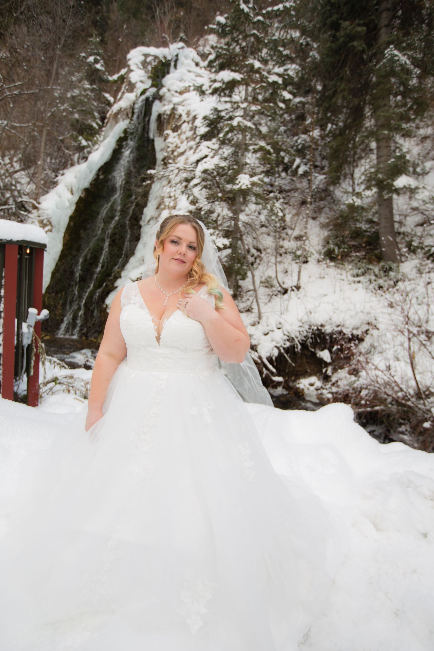 Elegant bride in white dress poses against frozen waterfall and snowy landscape.