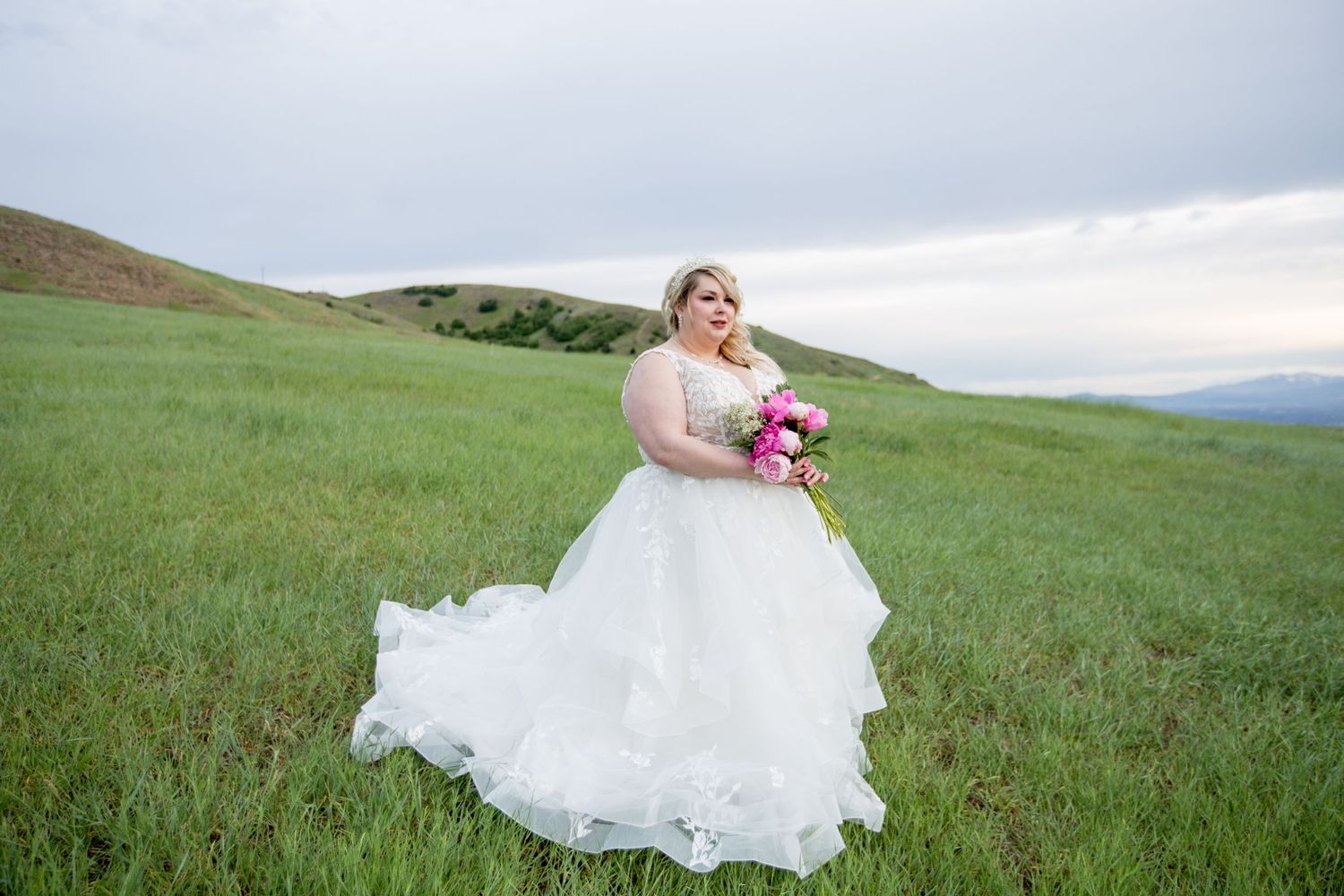 White wedding dress flows elegantly across rolling green hills under cloudy skies.