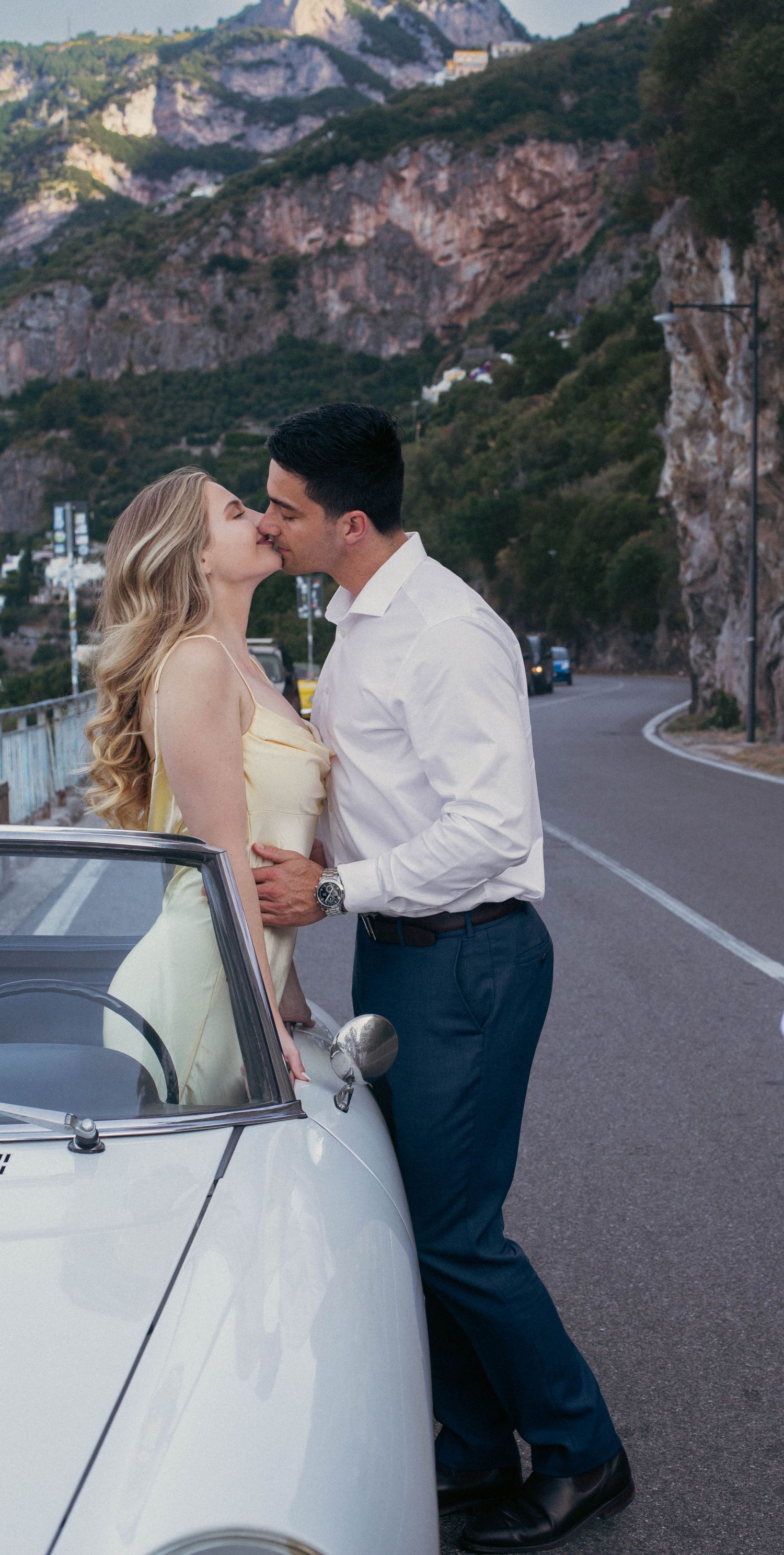 Couple sharing a kiss beside a vintage white convertible on a winding Amalfi Coast mountain road.