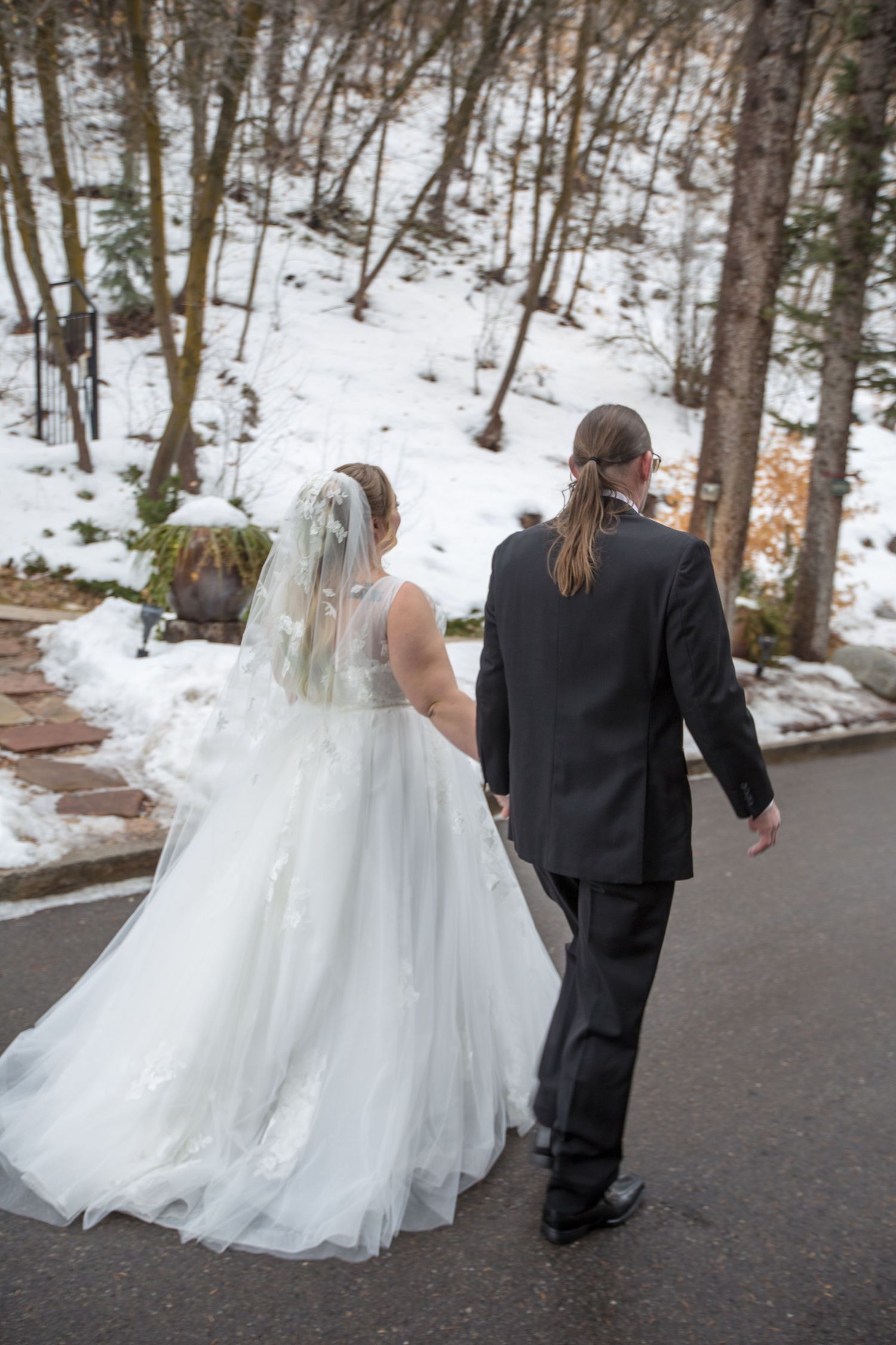 Bride in white gown with veil and groom in black suit walk hand in hand along snowy winter wedding venue path.