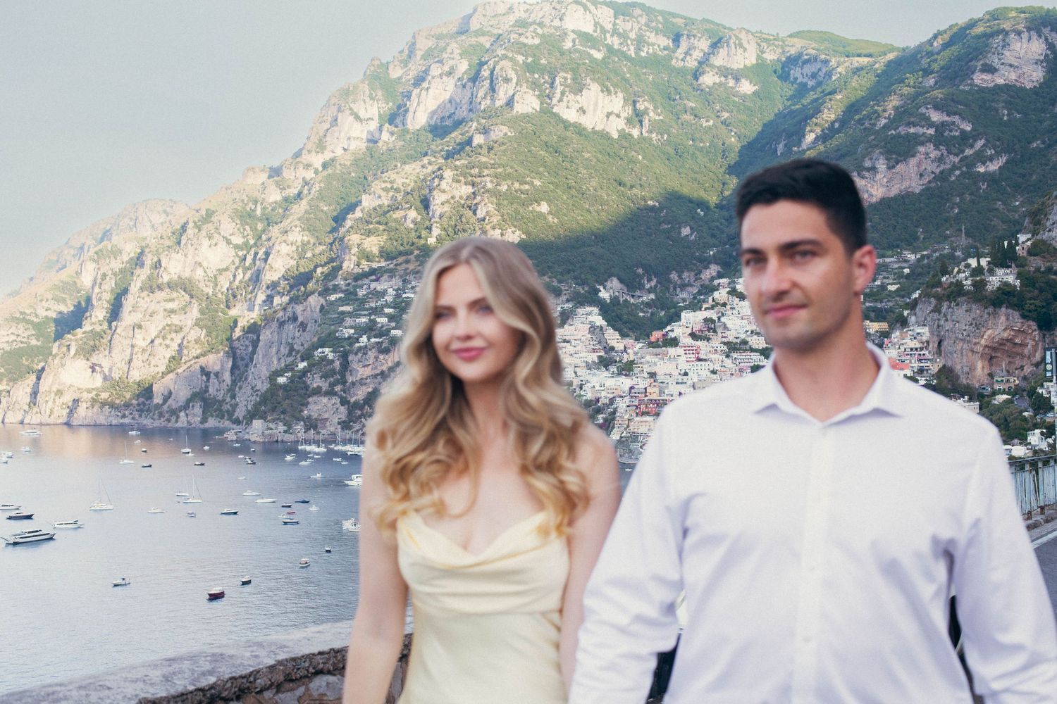Couple posing with Positano cliffside village and Mediterranean Sea in background, woman in yellow dress.