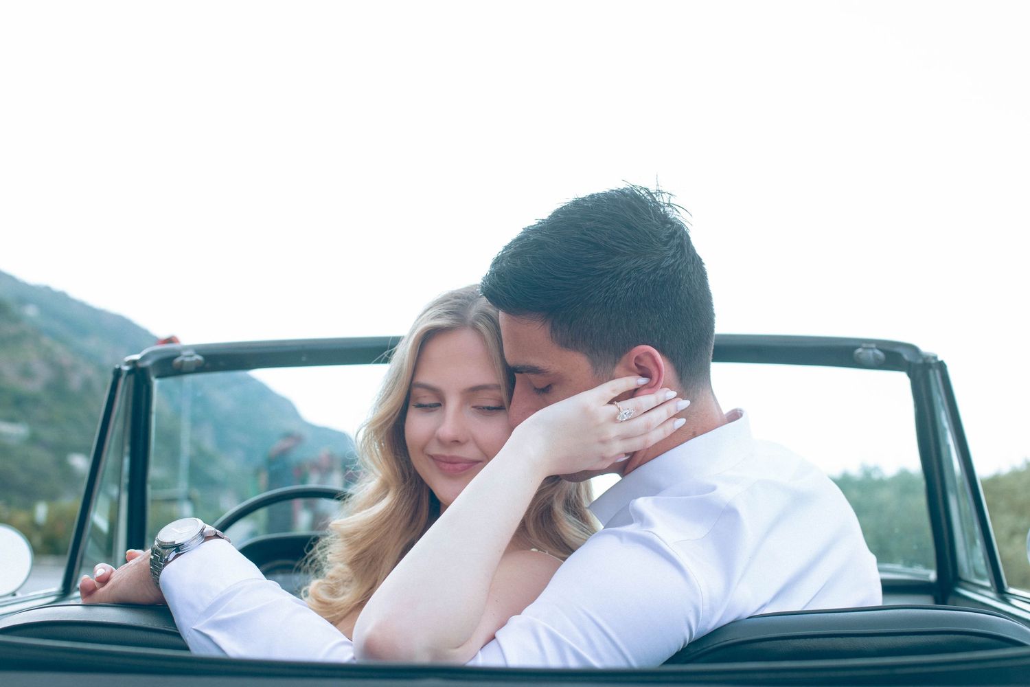 Woman embracing man tenderly in a vintage white convertible with mountains visible in the background.