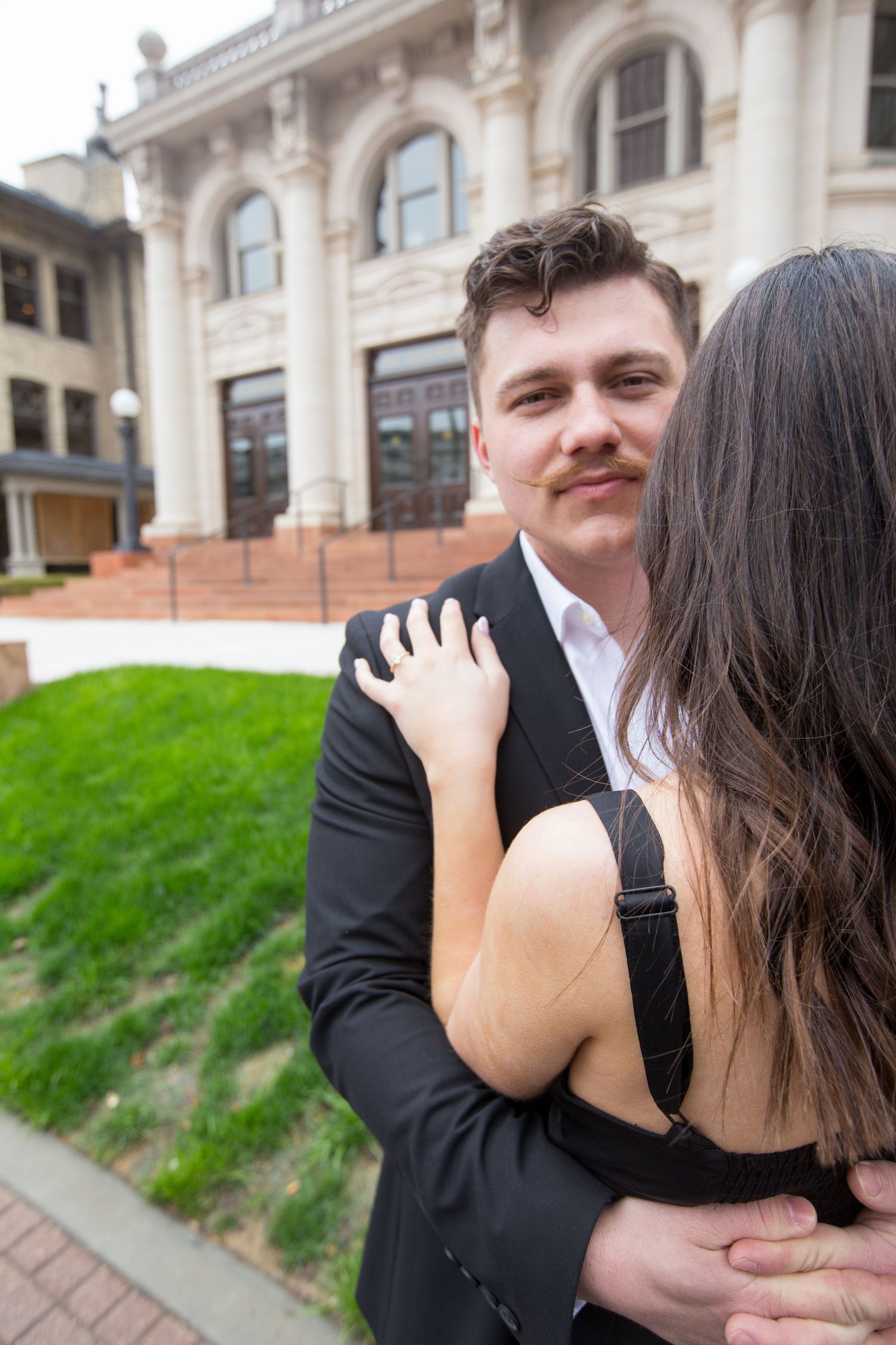 Couple embracing outside historic building, man in black suit with mustache smiling.