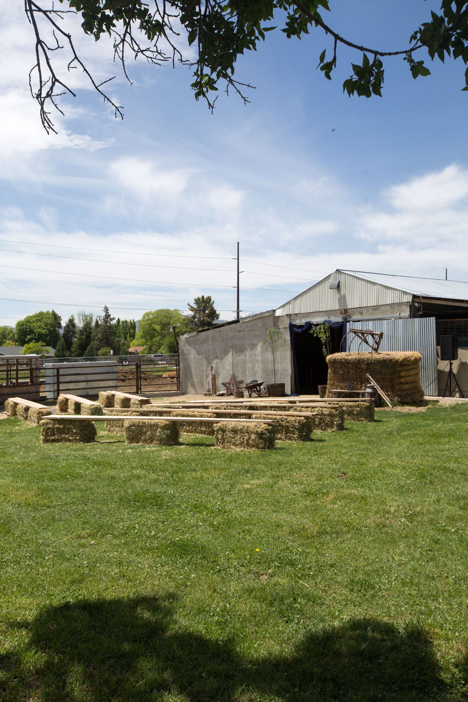 Farm yard with hay bales arranged on green grass beside a metal barn under a partly cloudy sky.