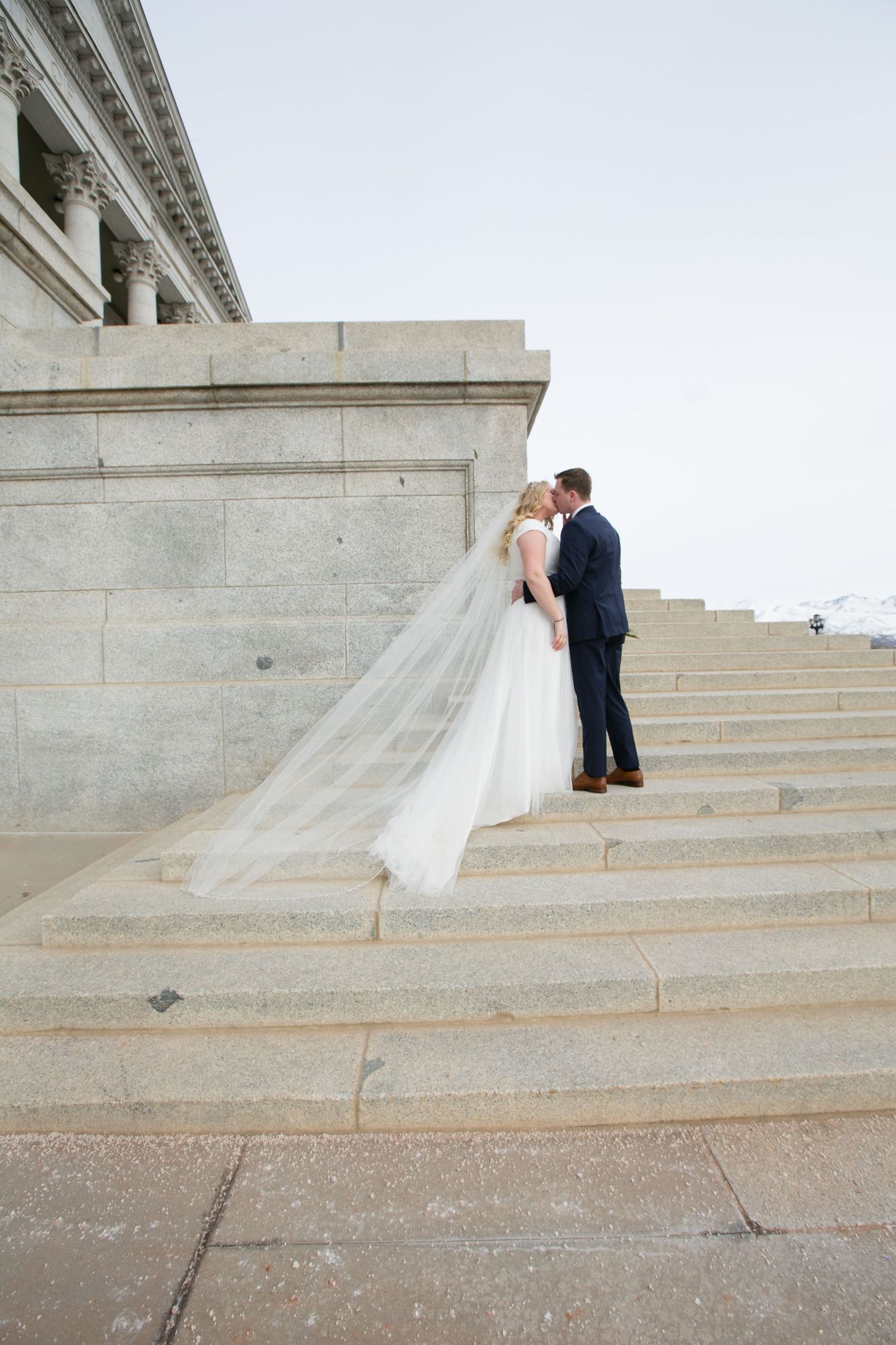 Bride and groom kissing on grand stone steps, bride's cathedral veil flowing, at a neoclassical building.