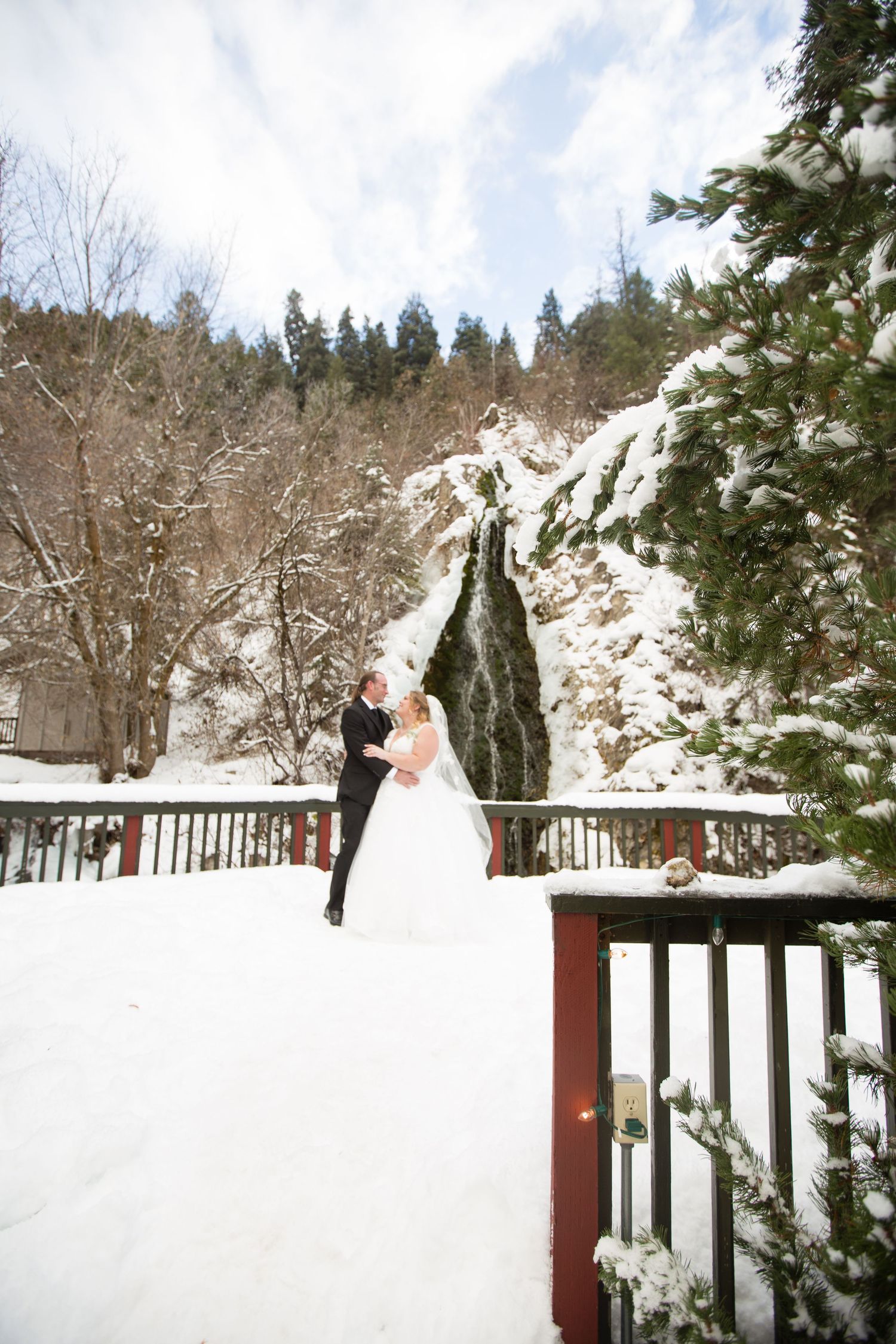 A winter wedding couple embraces on a snowy deck surrounded by evergreen trees and snow-covered mountains.