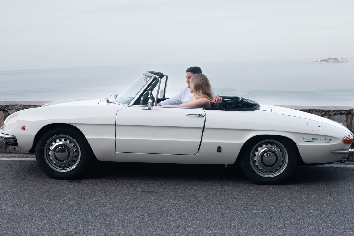 Couple kissing in white classic Alfa Romeo Spider convertible parked along the Amalfi Coast road.