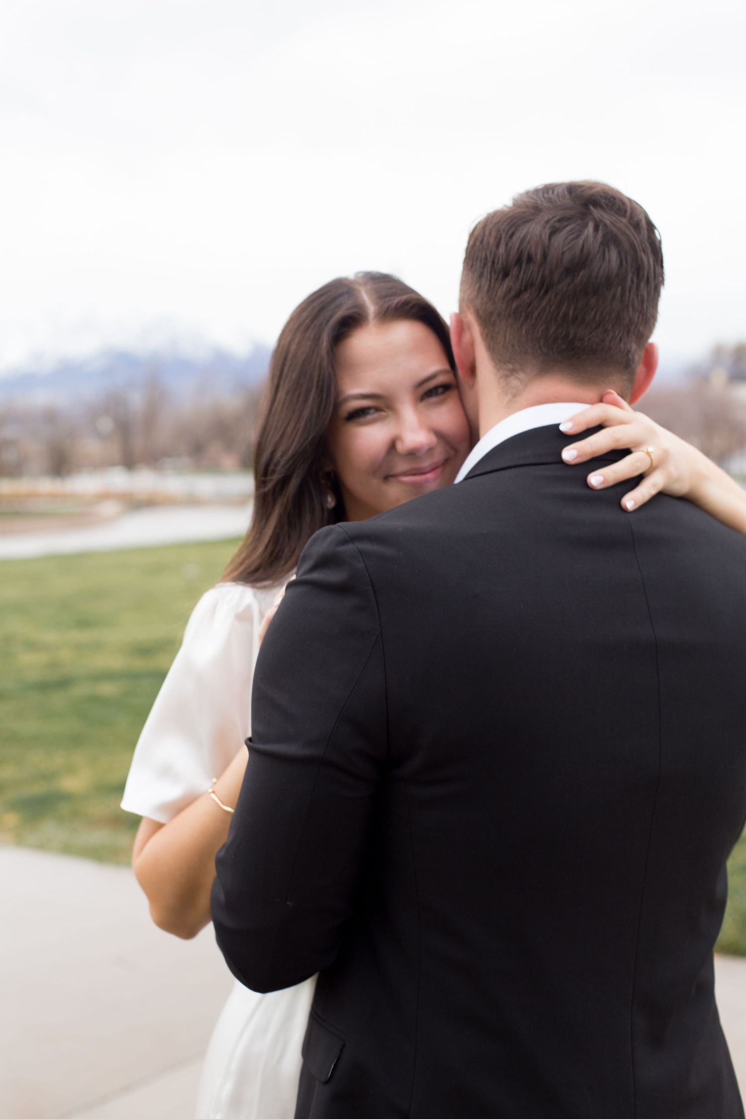 A bride in white dress smiles over groom's shoulder while embracing outdoors with mountains in background.